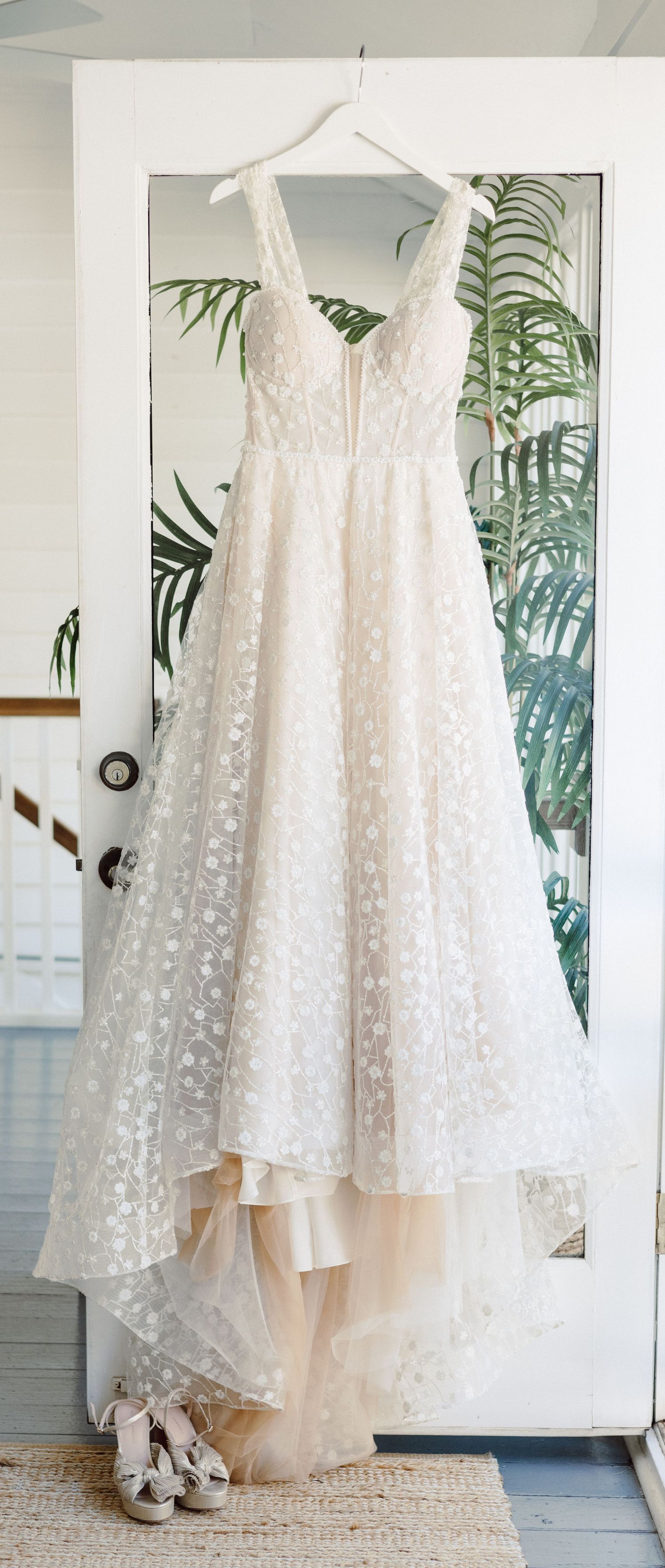 Delicate white lace wedding dress with floral details hanging against tropical palm leaf wallpaper.