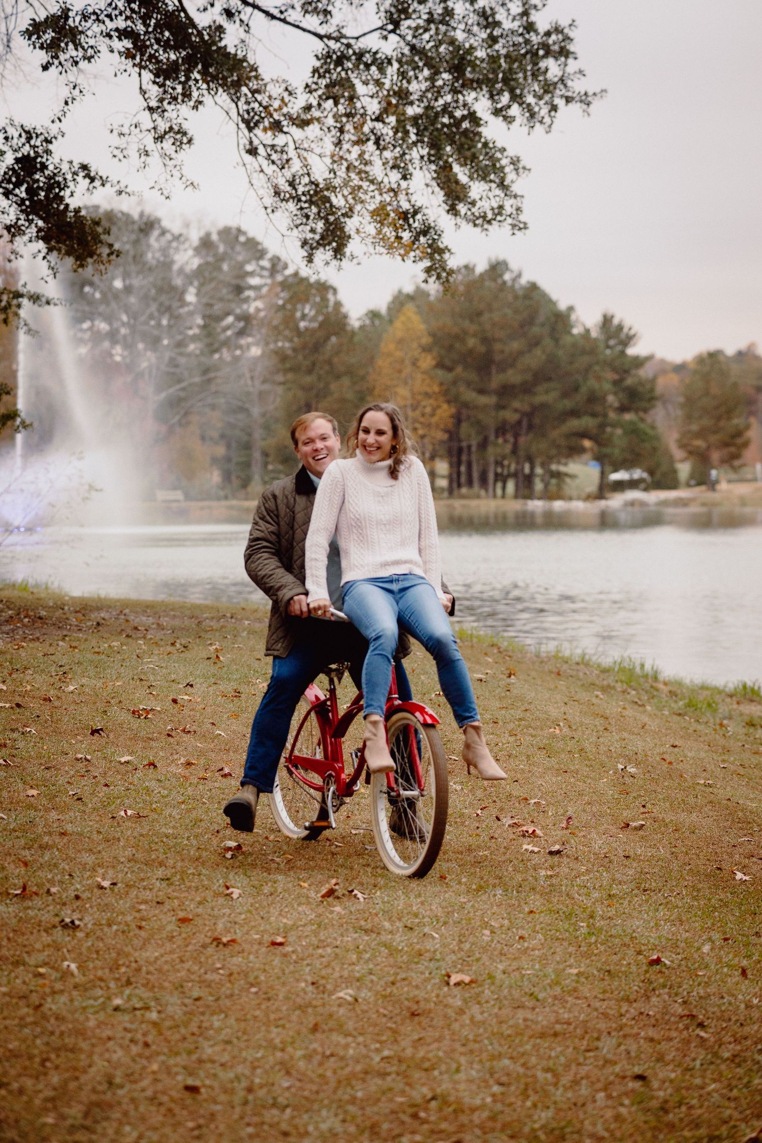 A couple shares a playful moment riding a bicycle together on an autumn day by a misty lake.