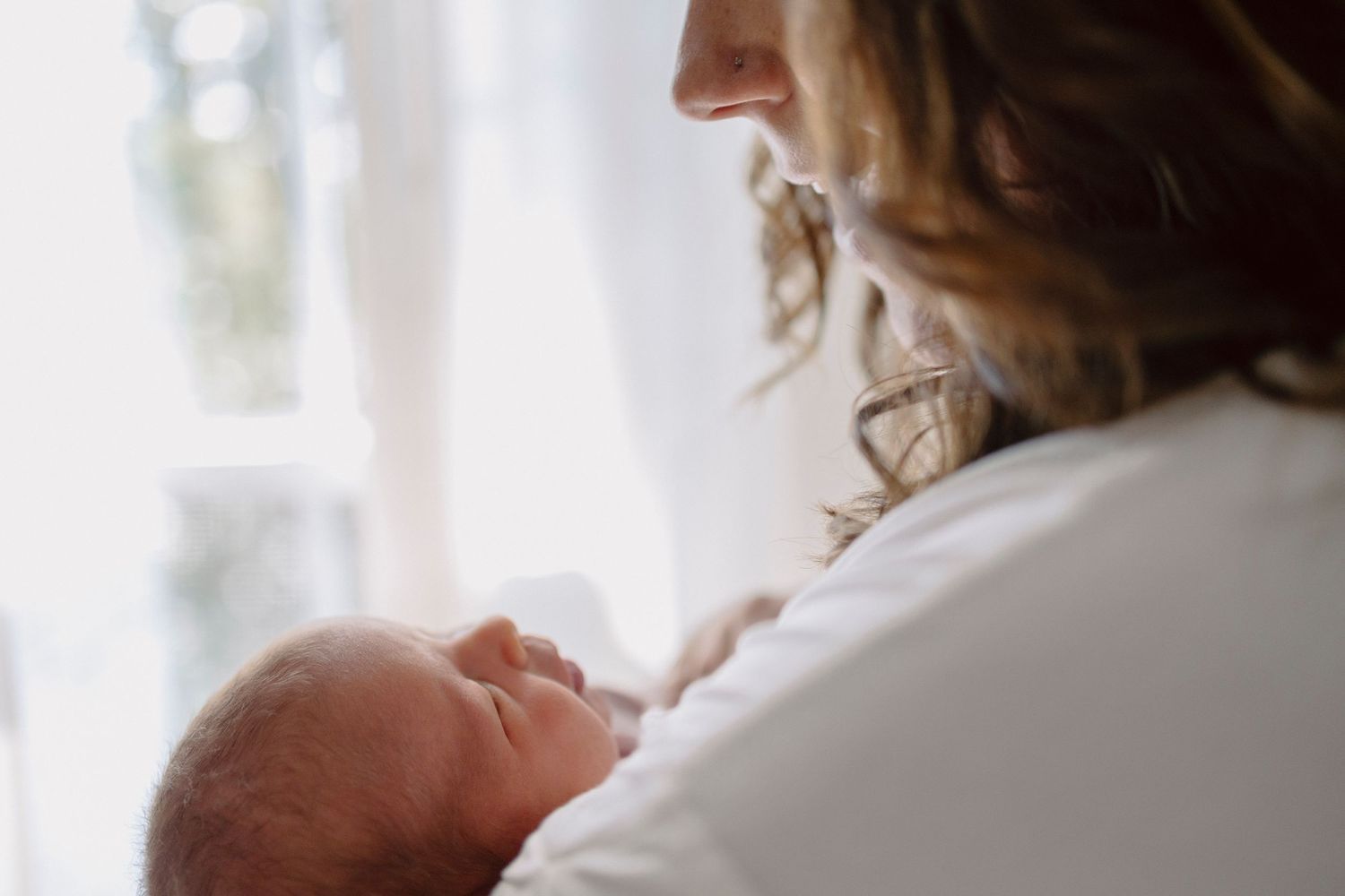 A person gently holds and gazes at a newborn baby near a sunlit window.