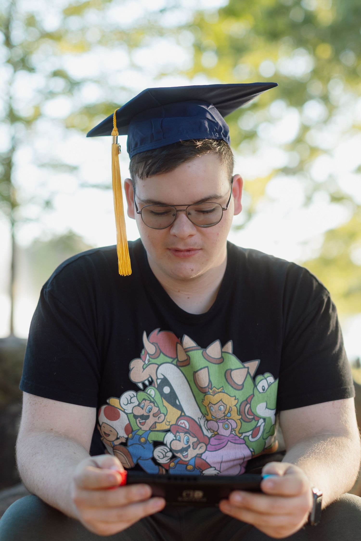 Young man wearing graduation cap and Mario t-shirt plays Nintendo Switch outdoors, celebrating academic achievement.