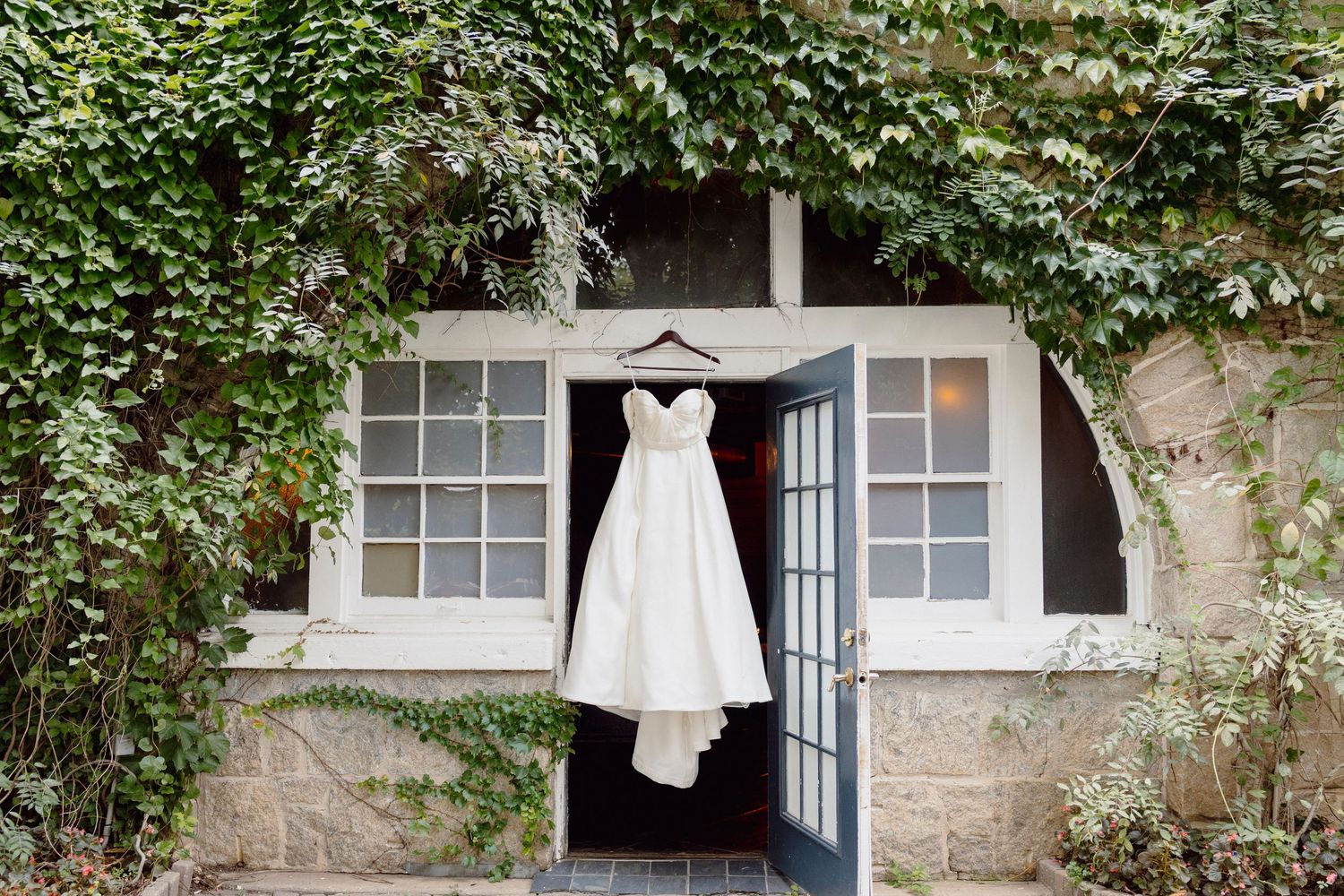 White wedding dress hanging in front of ivy-covered stone building exterior.