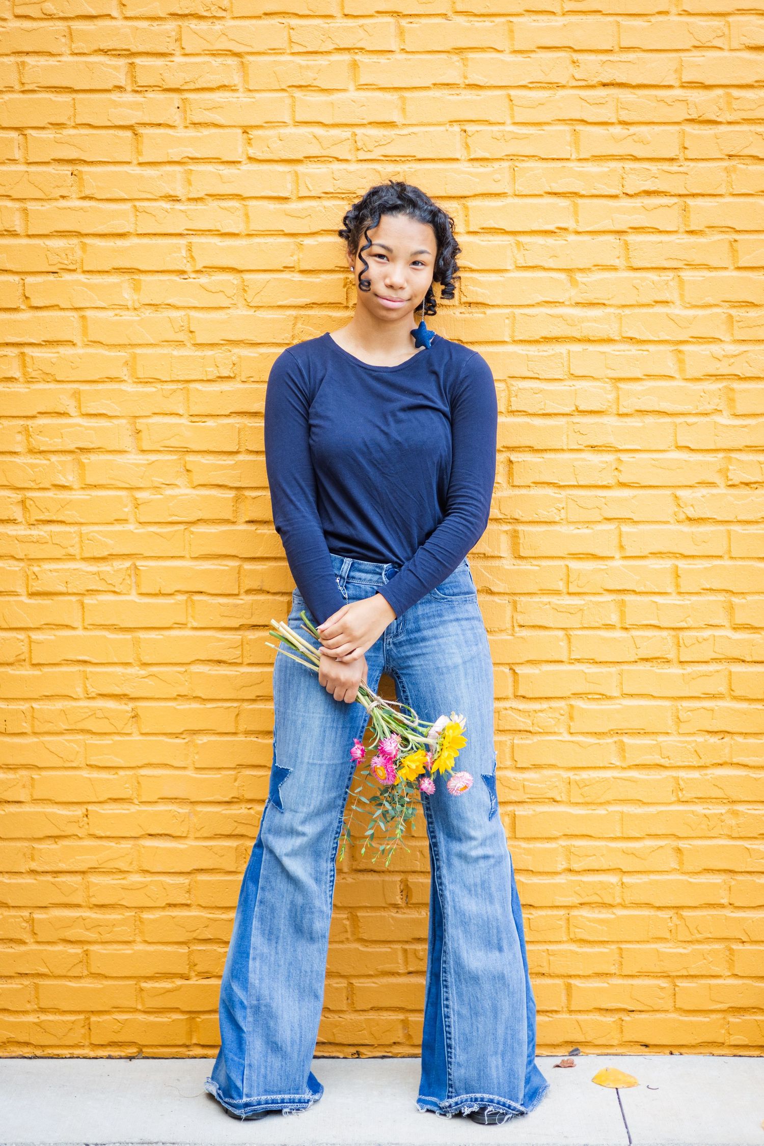 Young Black woman in navy top and wide-leg jeans holds colorful wildflowers against a bright yellow brick wall.
