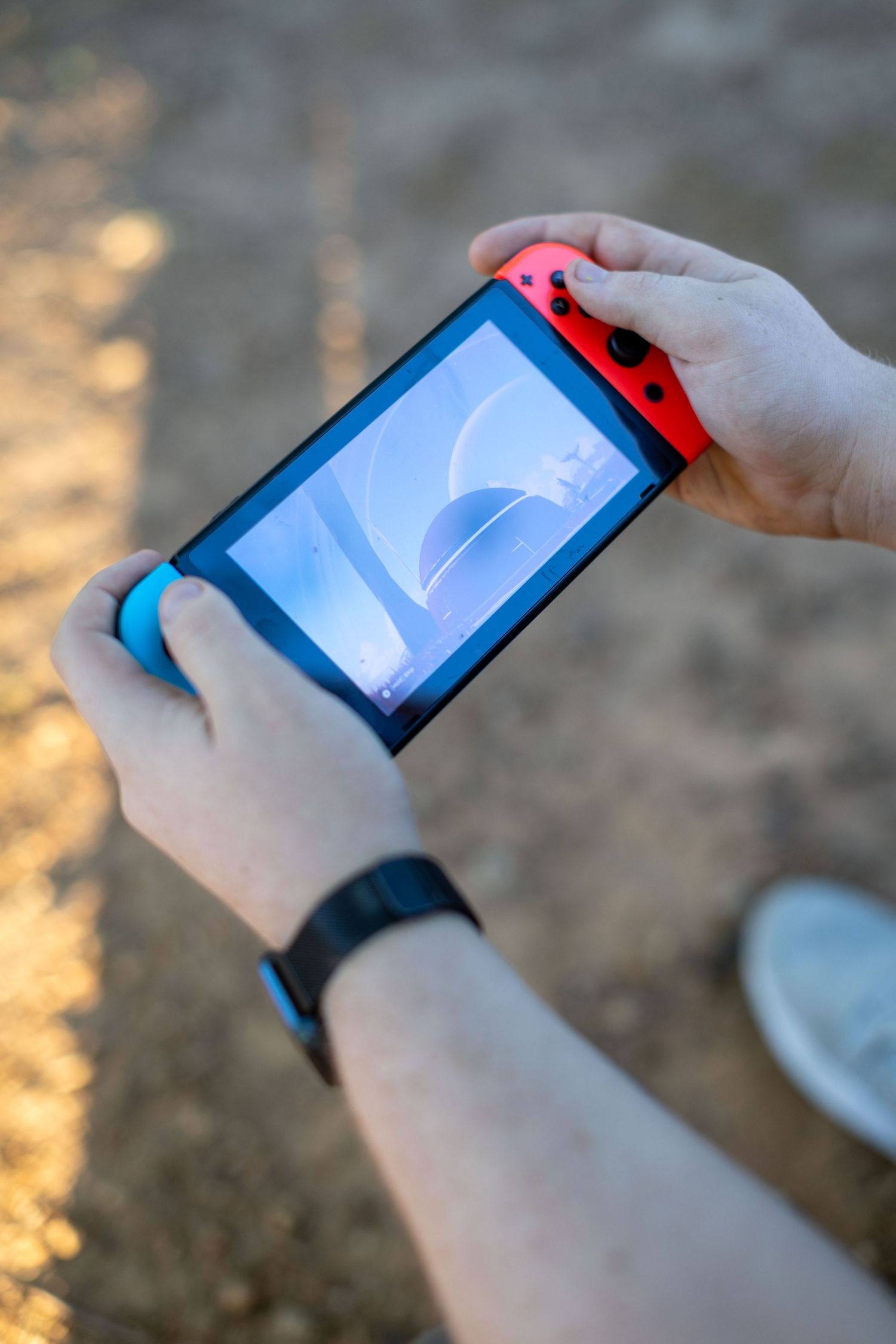 Person holding a Nintendo Switch console with red and blue Joy-Con controllers outdoors in handheld mode.