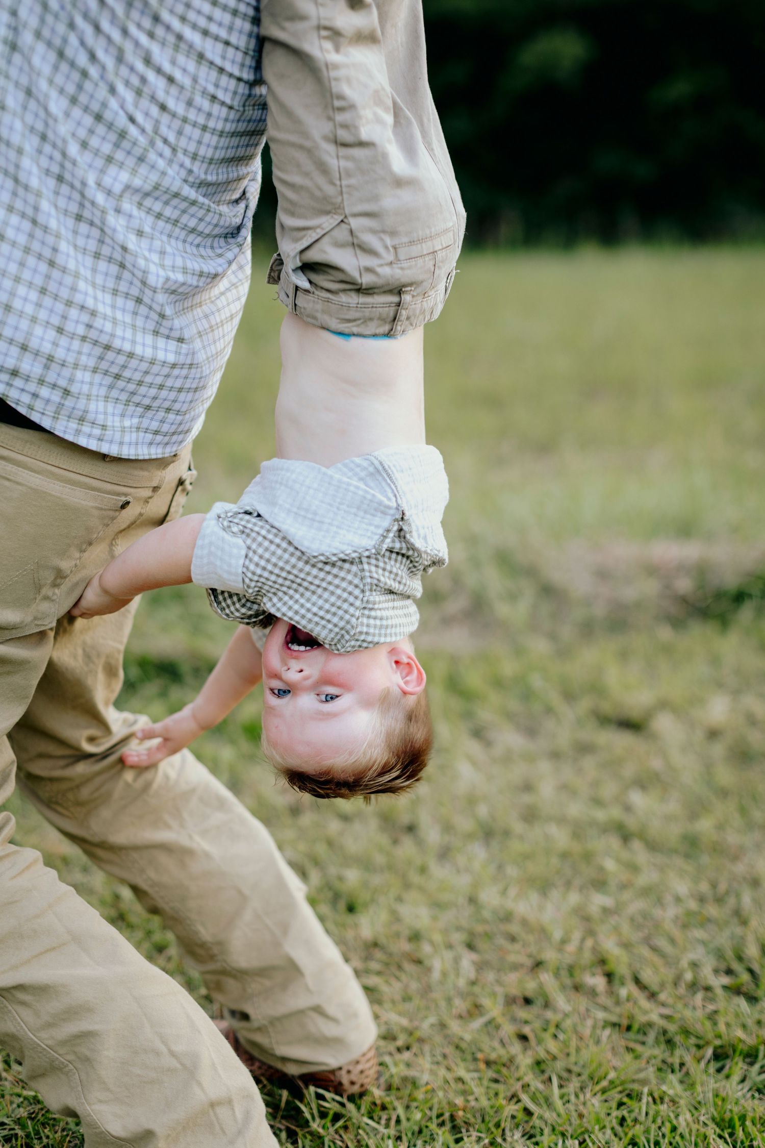 Father playfully holding young child upside down outdoors on green grass field during fun family playtime.