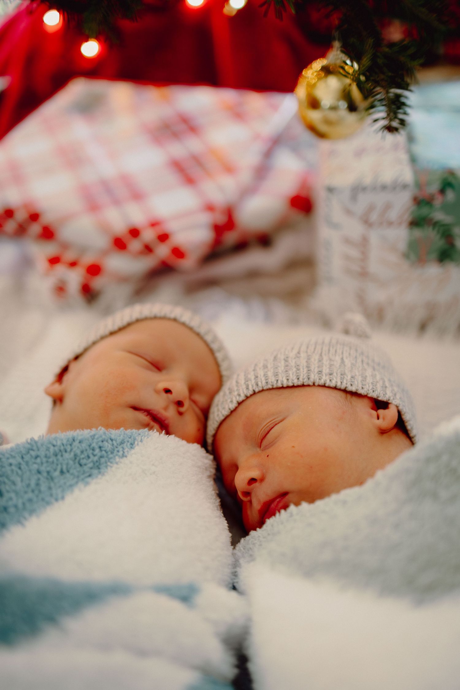 Sleeping baby wrapped in white knit blanket under soft red Christmas lights and holiday decorations.