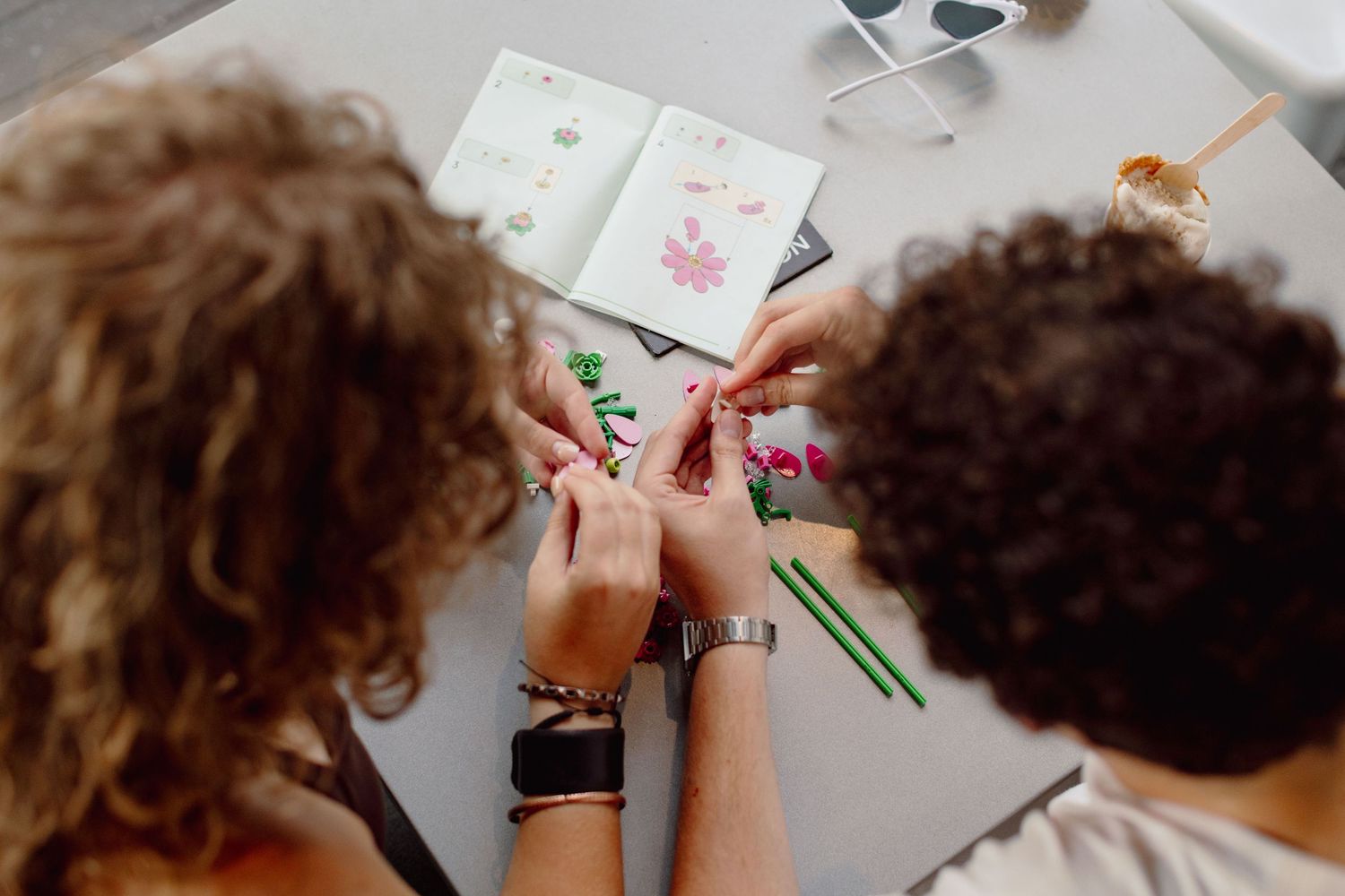 Two people collaborate on a colorful craft project, arranging small decorative elements on a table with cards nearby.