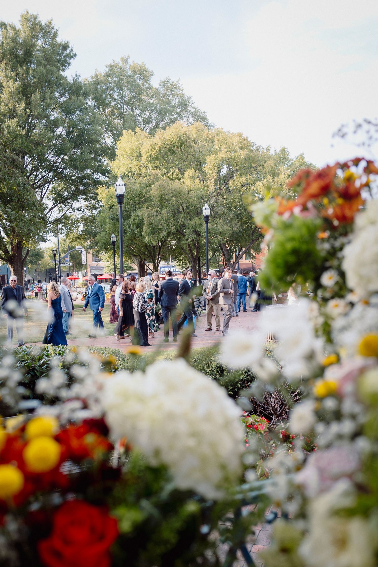 Colorful flowers in the foreground frame a blurred crowd of people walking along a tree-lined path in a park.