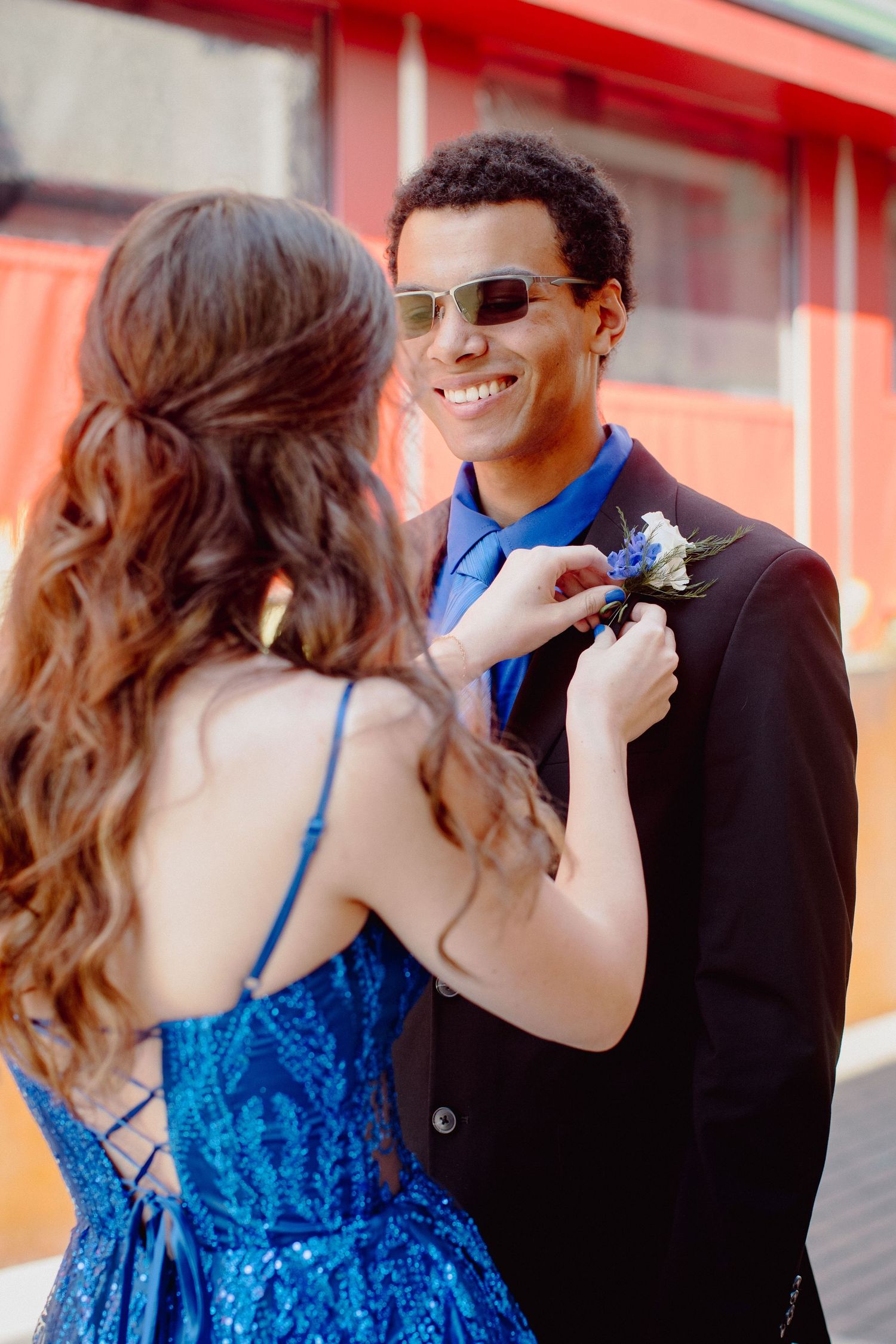 A young woman in a blue dress pins a boutonniere on a smiling man in a black suit at prom.