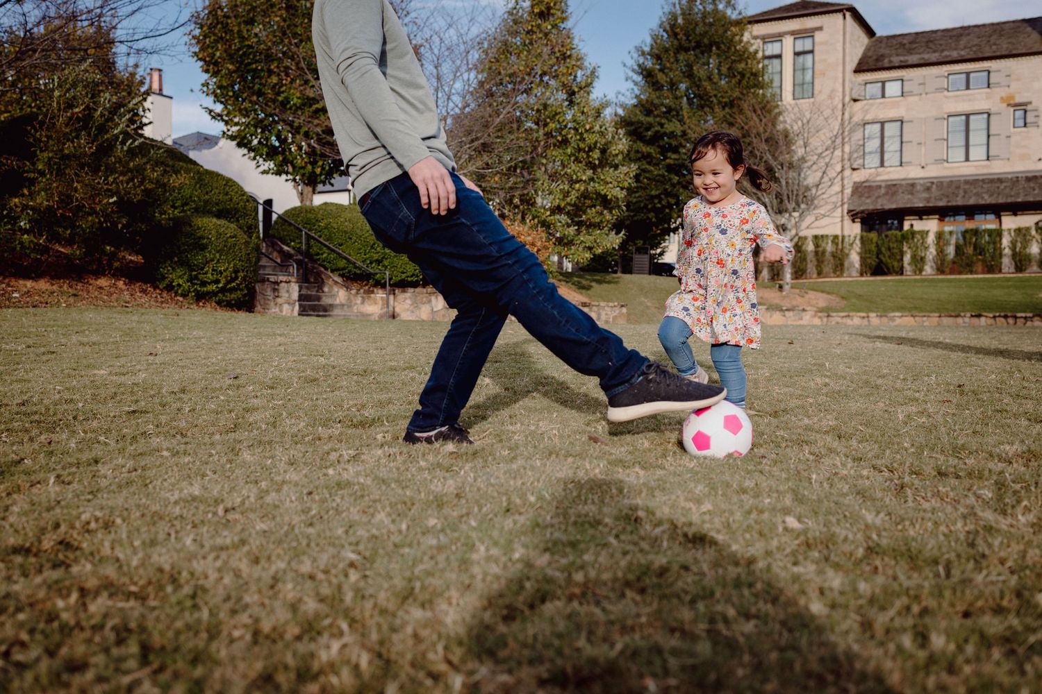 Two people play soccer together on a grassy lawn outside a brick building on an autumn day.