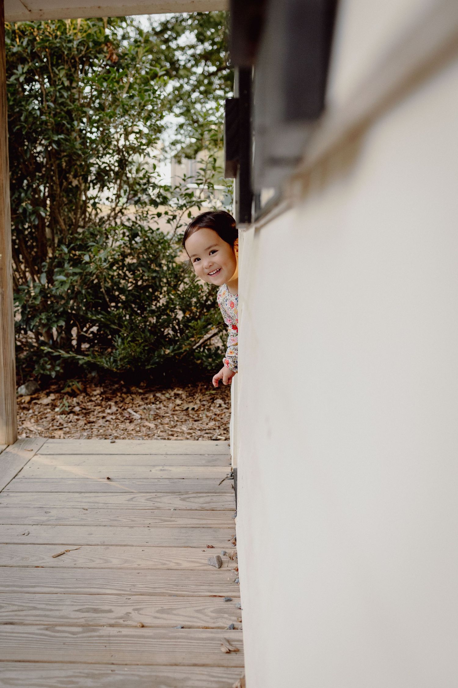 Someone playfully peeks around a white wall on a wooden deck with greenery in the background.