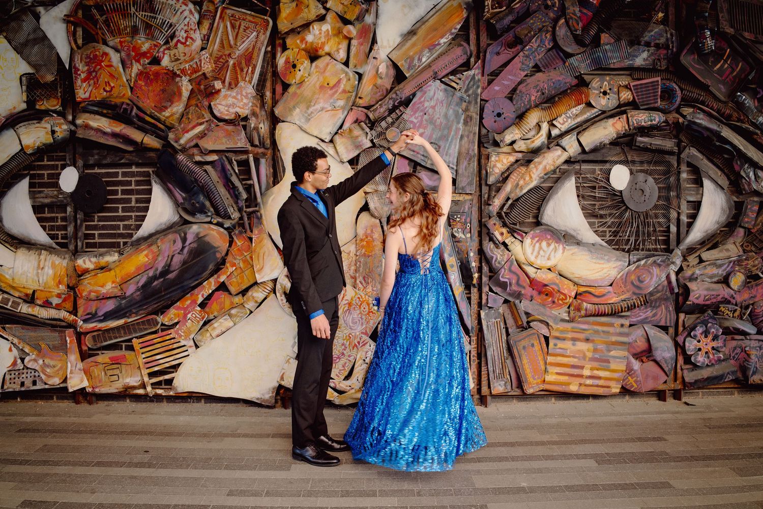 Couple dancing in formal wear before a large colorful graffiti mural featuring giant eyes.
