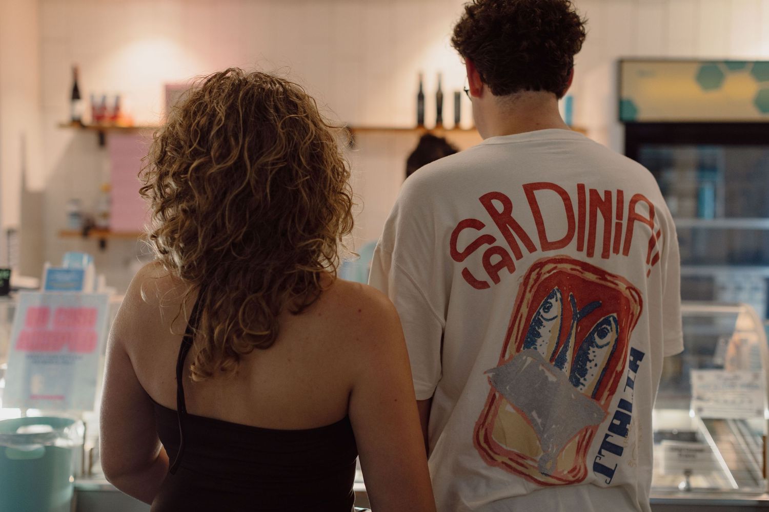 Two people stand at a counter, one wearing a white graphic Sardinifed t-shirt in a casual cafe setting.