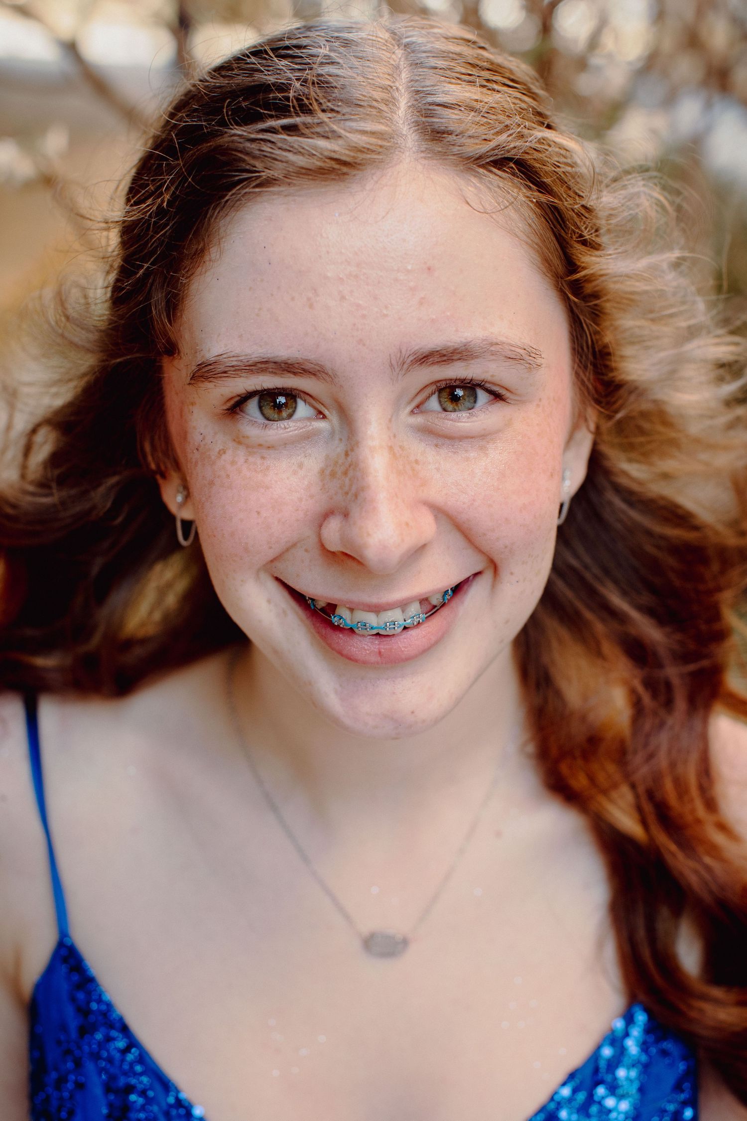 Smiling teenage girl with braces, freckles, and brown hair wearing a blue sequin dress and delicate necklace outdoors.