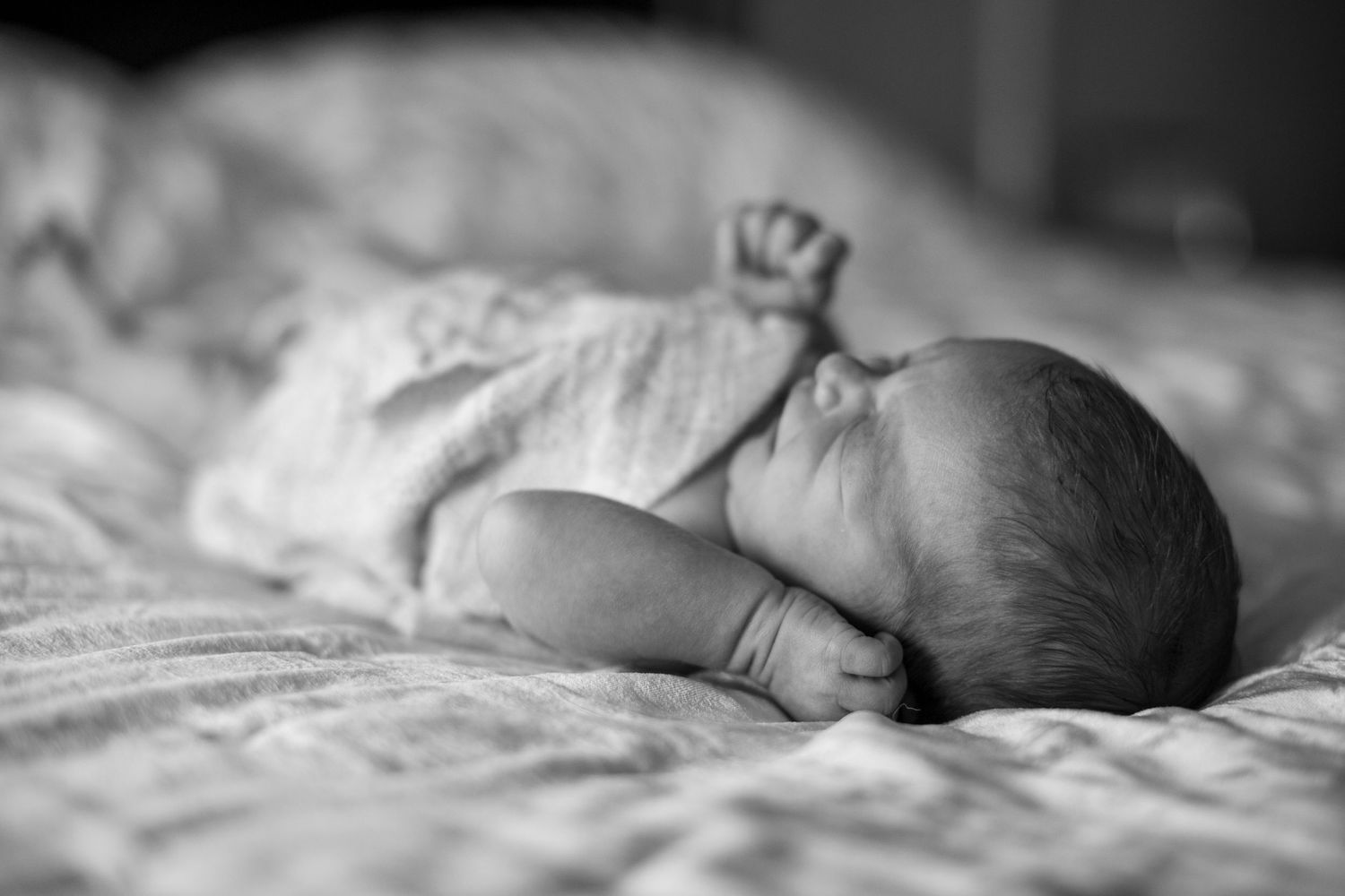 Black and white photo of a peaceful newborn baby sleeping on a soft bed, wrapped in a white swaddle blanket.