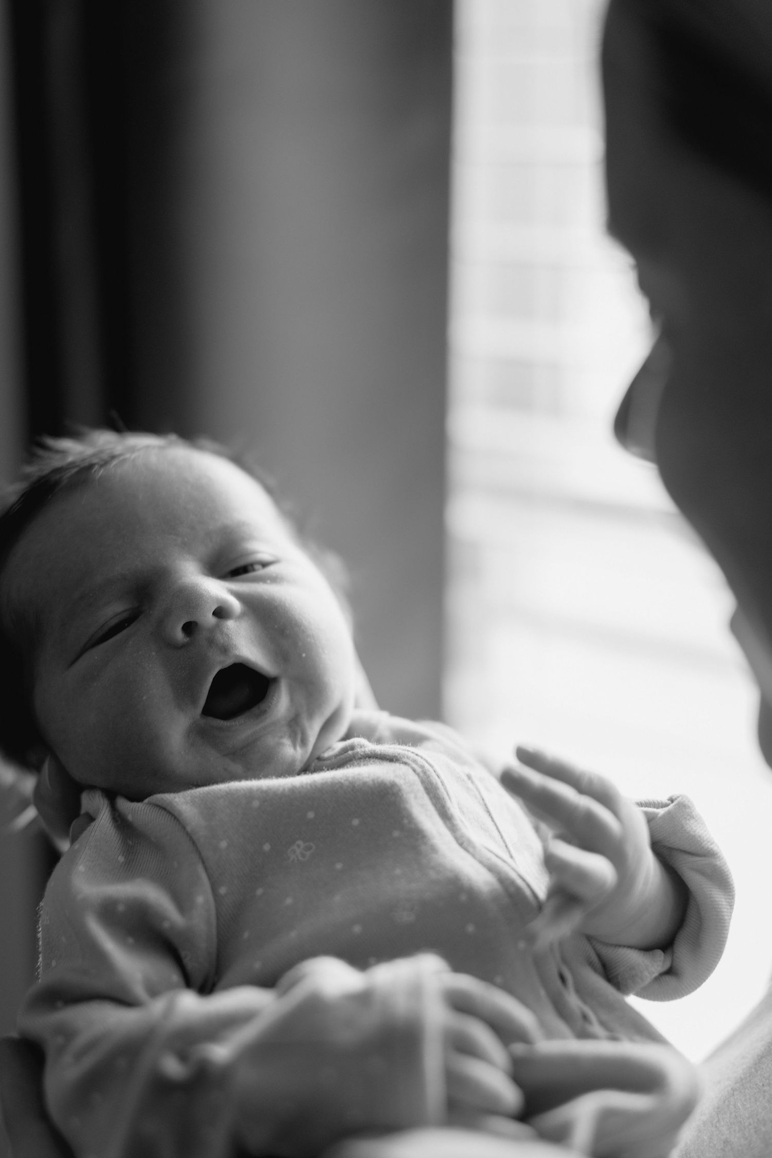 Black and white photo of a yawning newborn baby being cradled in loving arms near a bright window.