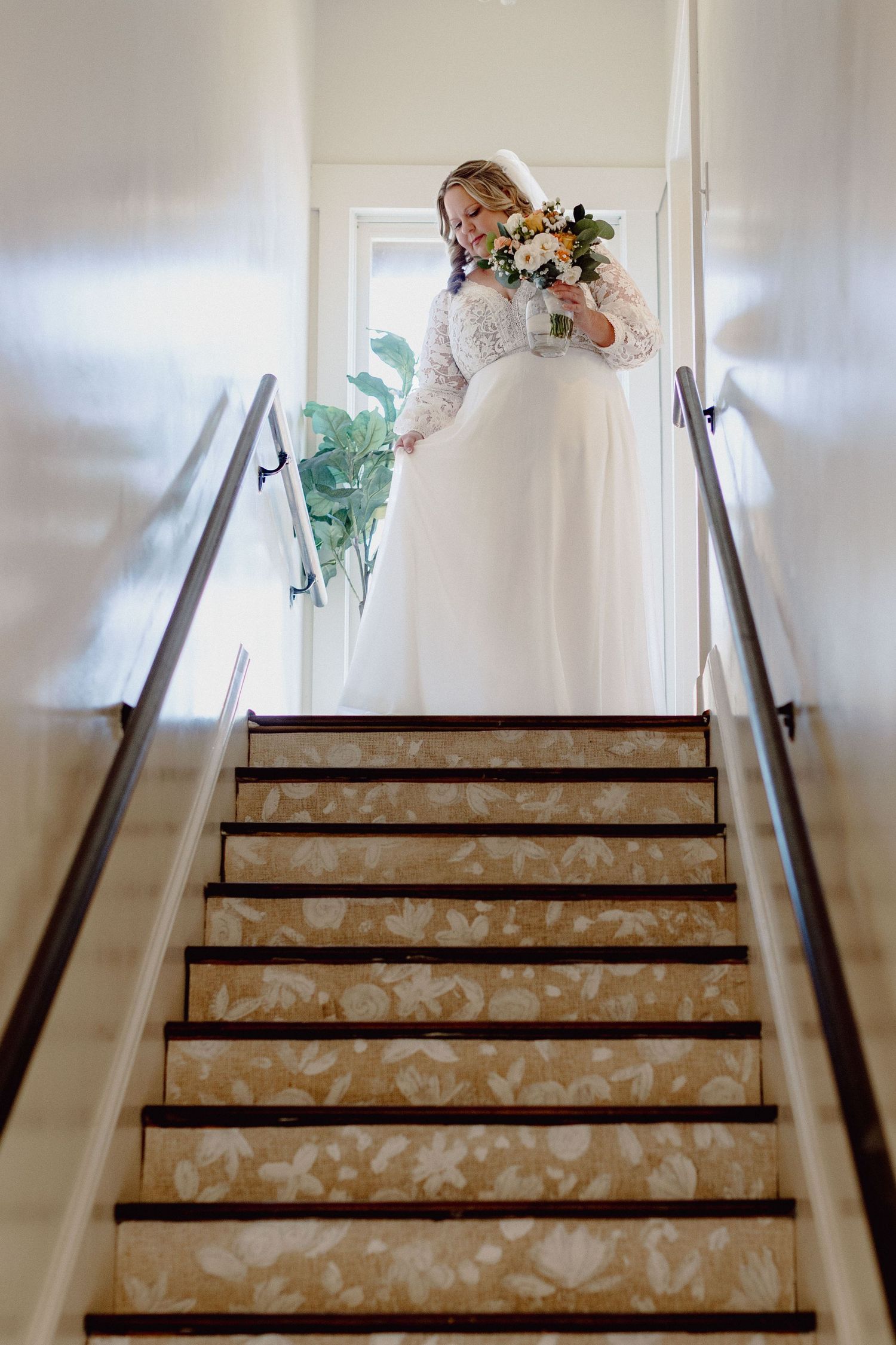 A bride in a white lace gown stands at the top of a staircase holding a bouquet of flowers.