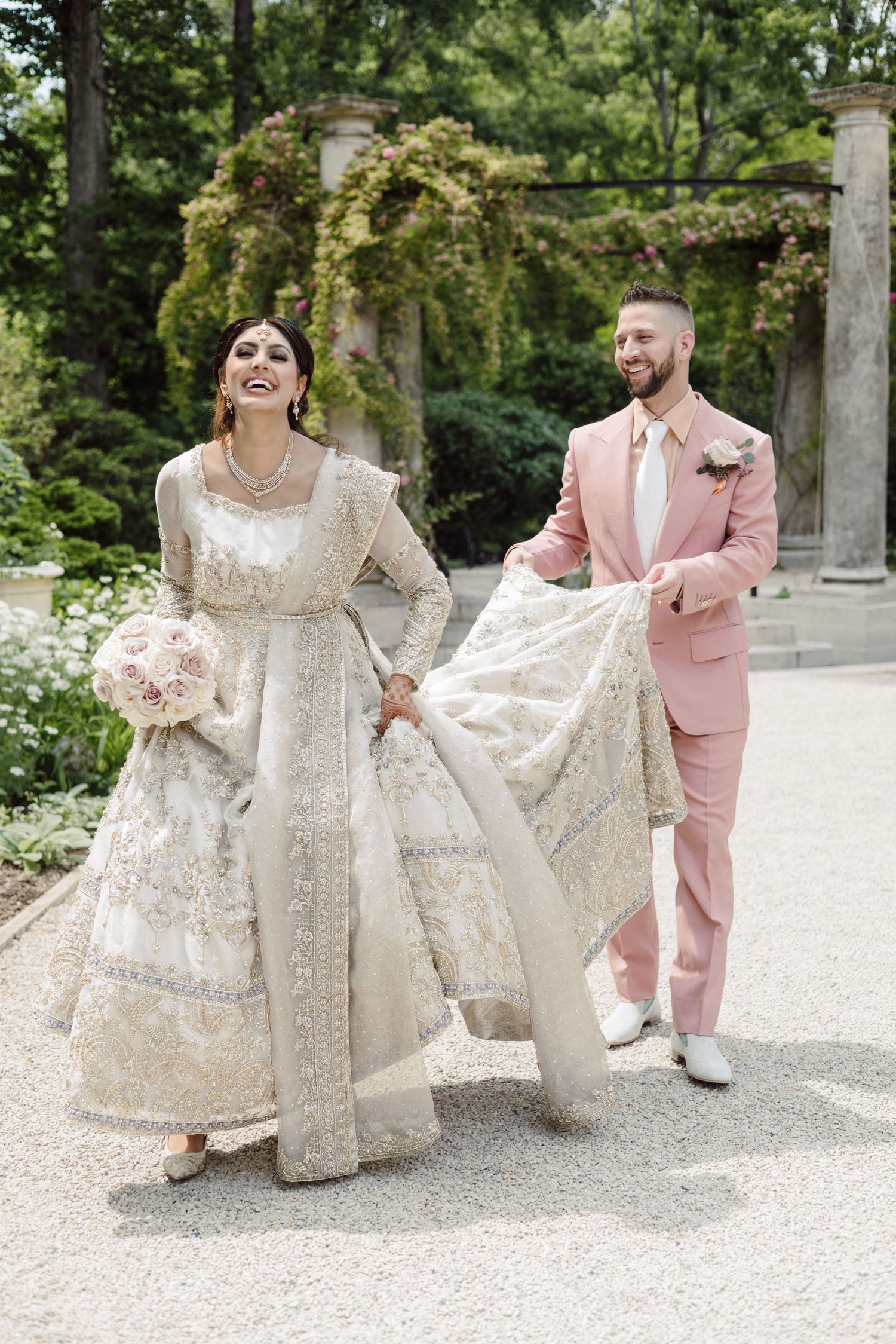 Couple in pink suit and white lace dress posing playfully on gravel path.