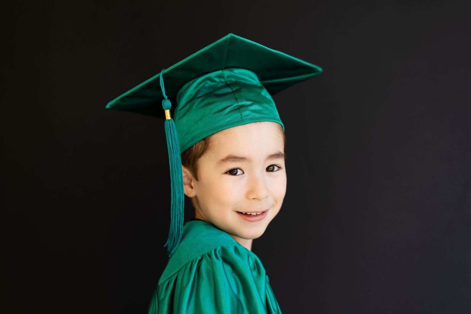 Young child smiling in green graduation cap and gown against dark background.