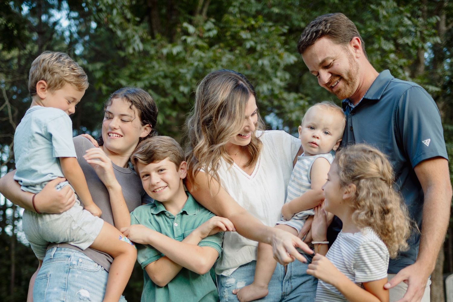 A family plays and laughs together during an outdoor photo session in a forest setting.