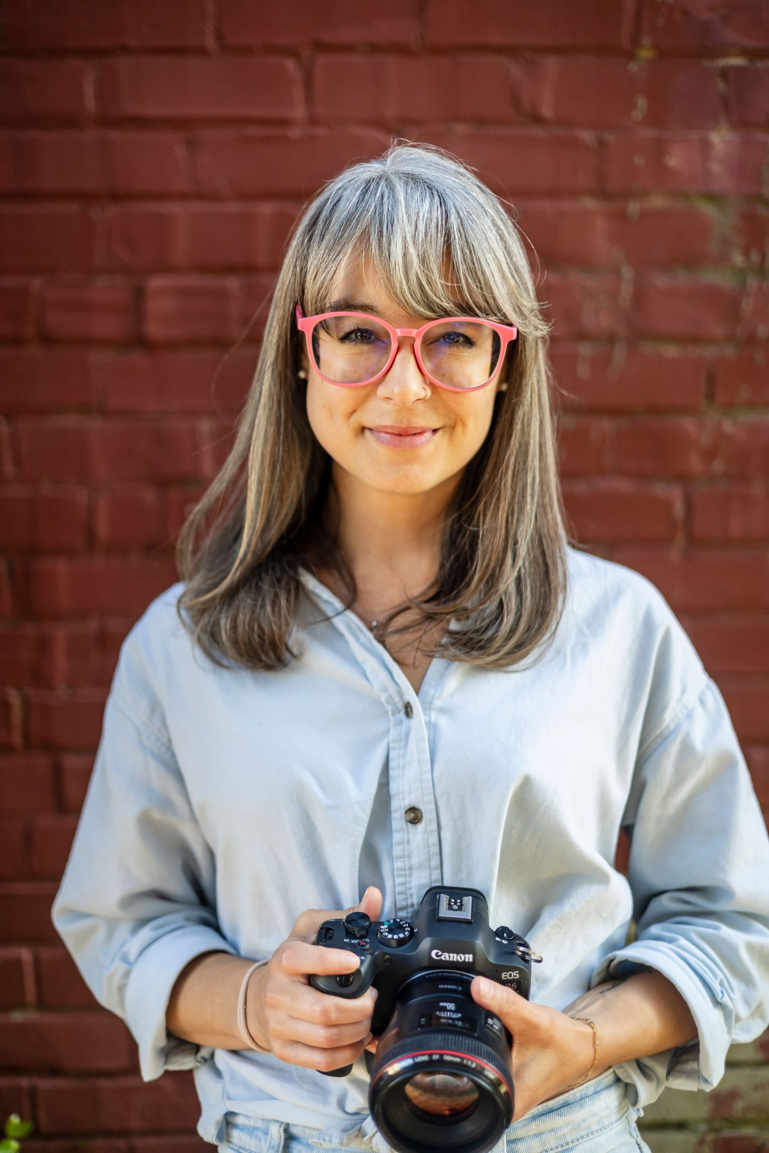 A photographer wearing pink glasses and a light gray sweater holds a professional camera against a brick wall background.