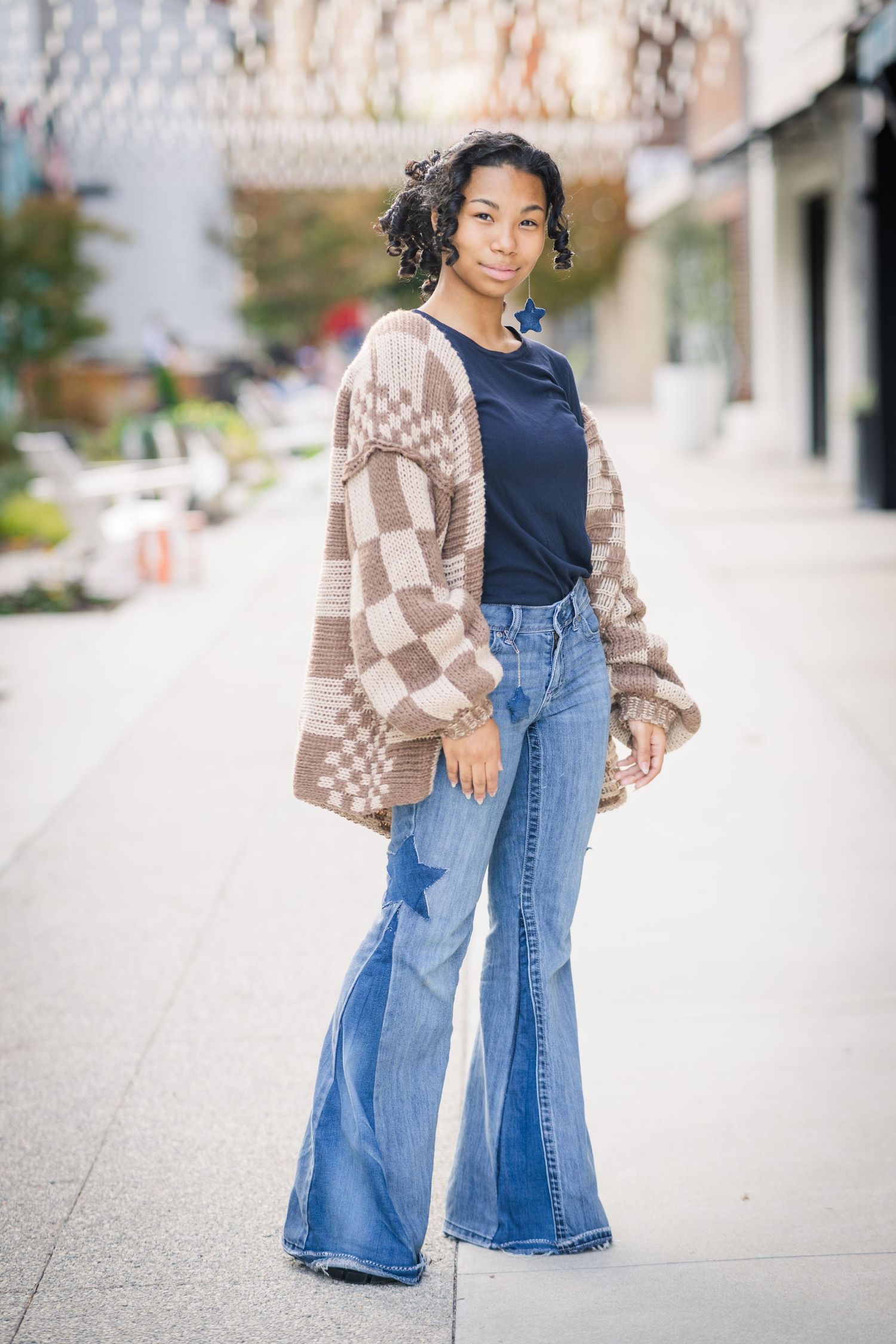 Young woman in flare jeans, navy top, and checkered cardigan posing on urban sidewalk.
