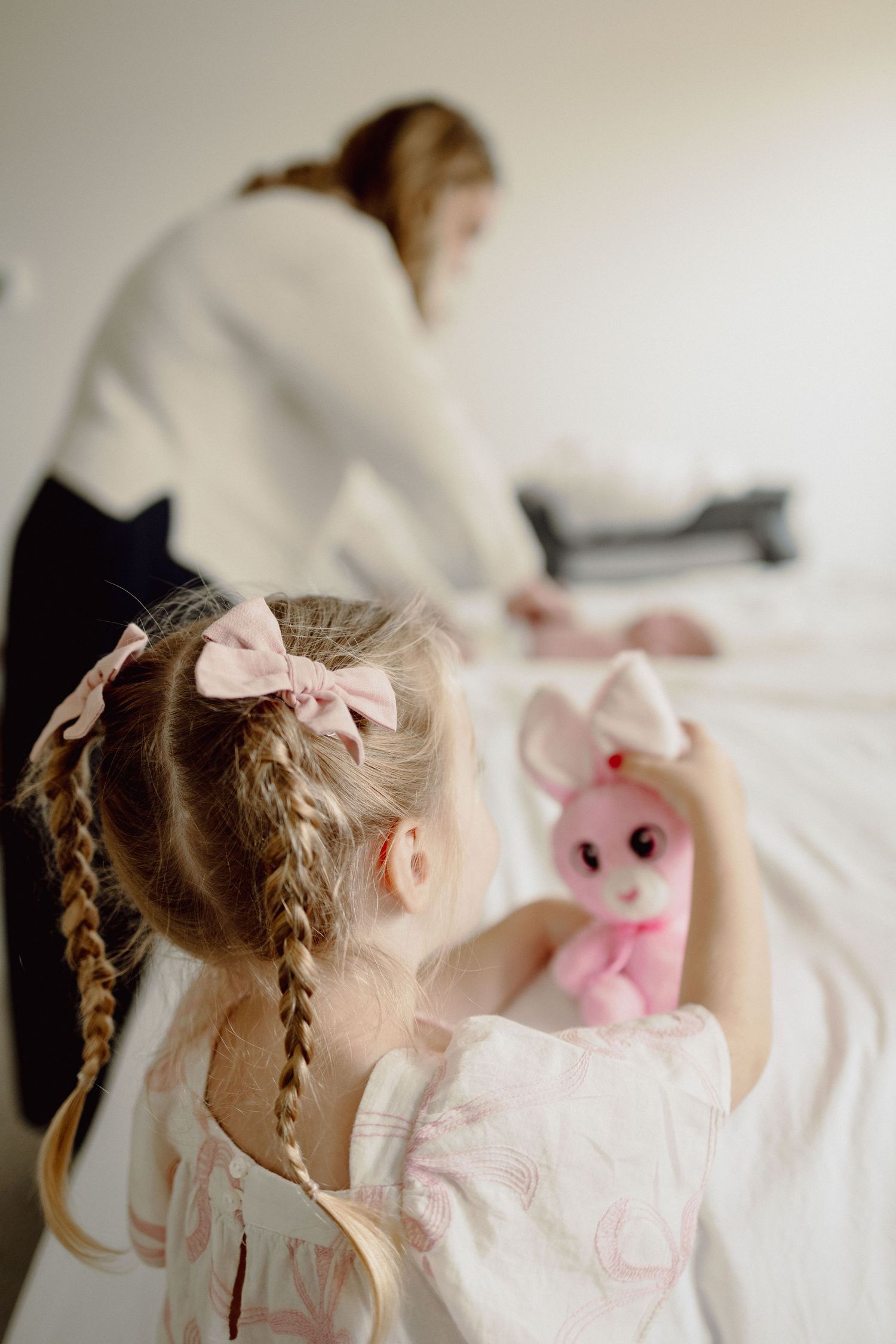 A young child in a pink dress cuddles with a plush pink bunny toy on white bedding.