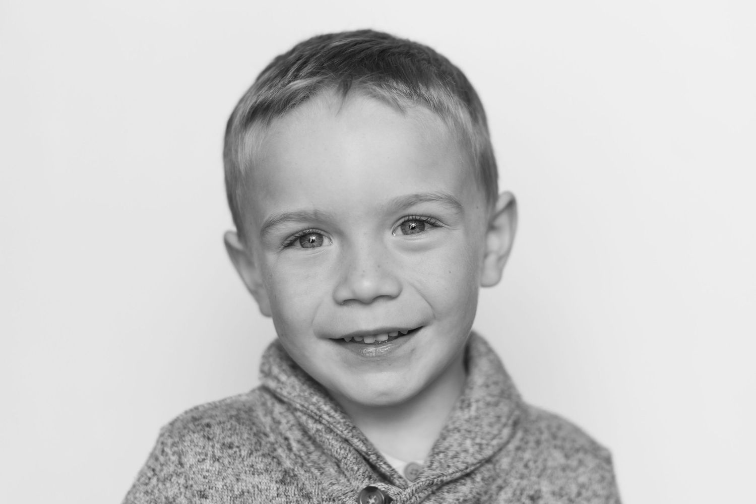 Black and white portrait of a smiling young blonde boy wearing a sweater against a light background.