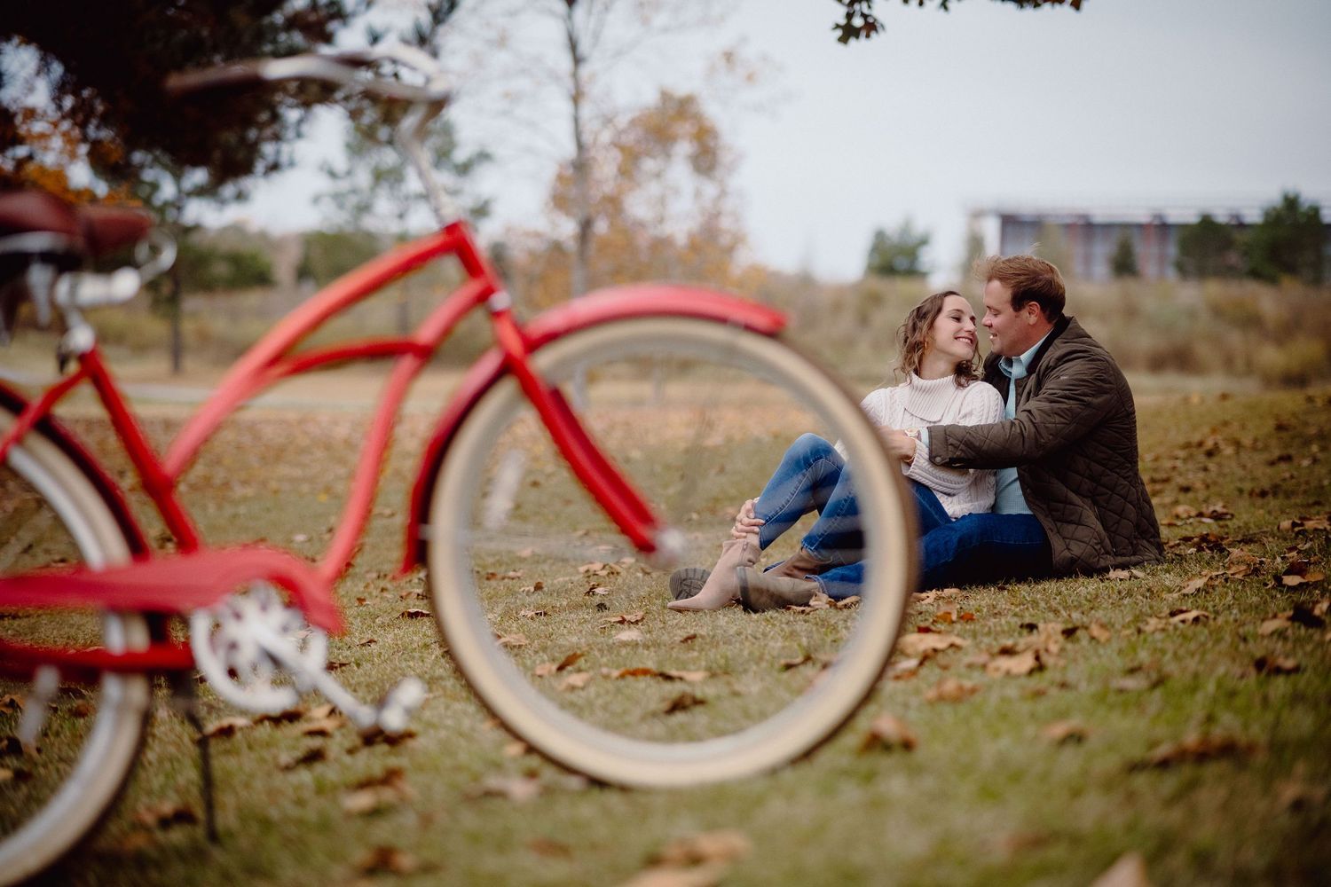 A romantic couple sits together on autumn ground near a vintage red bicycle in a park setting.