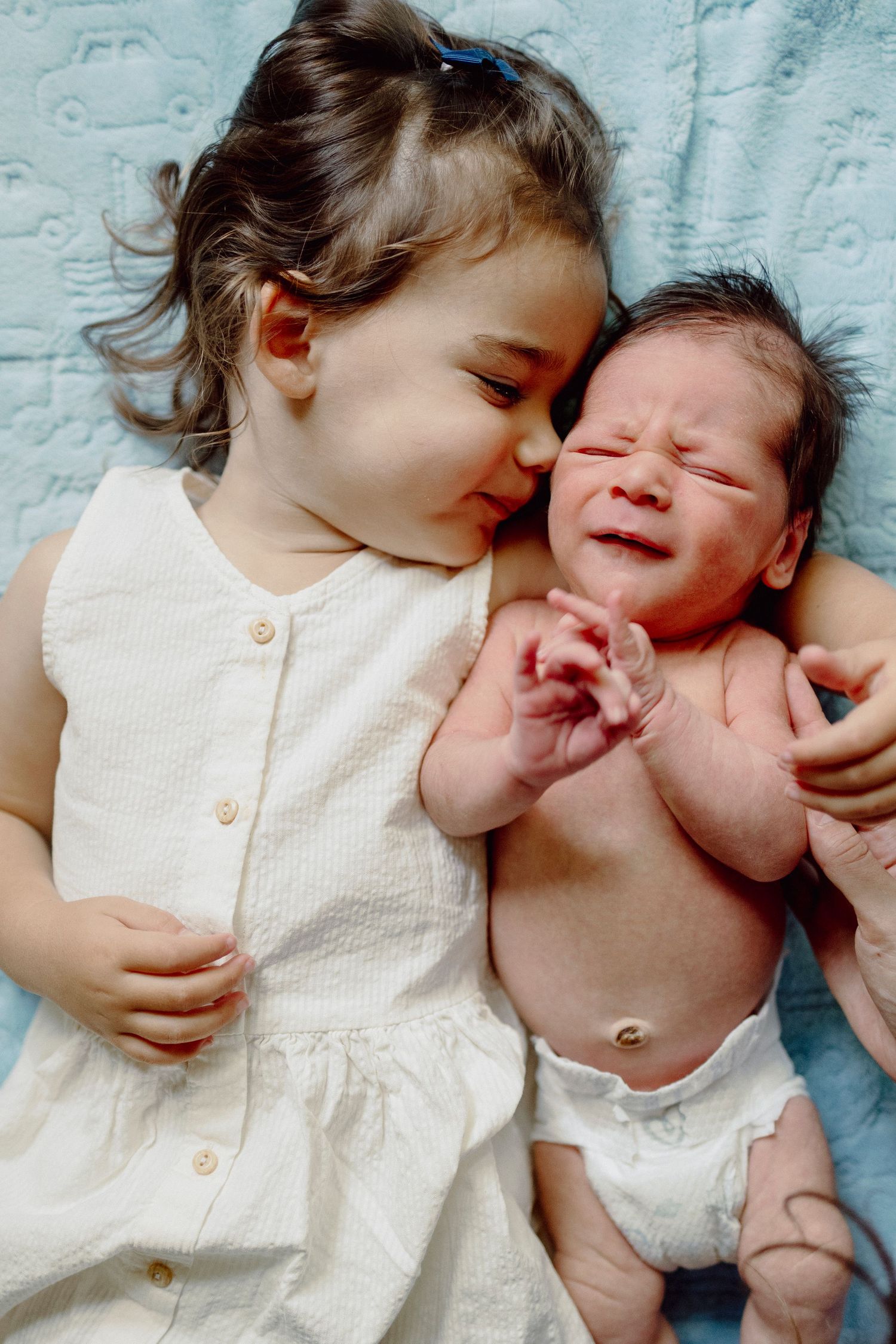 A toddler in a white dress lies next to a crying newborn baby on a light blue blanket.
