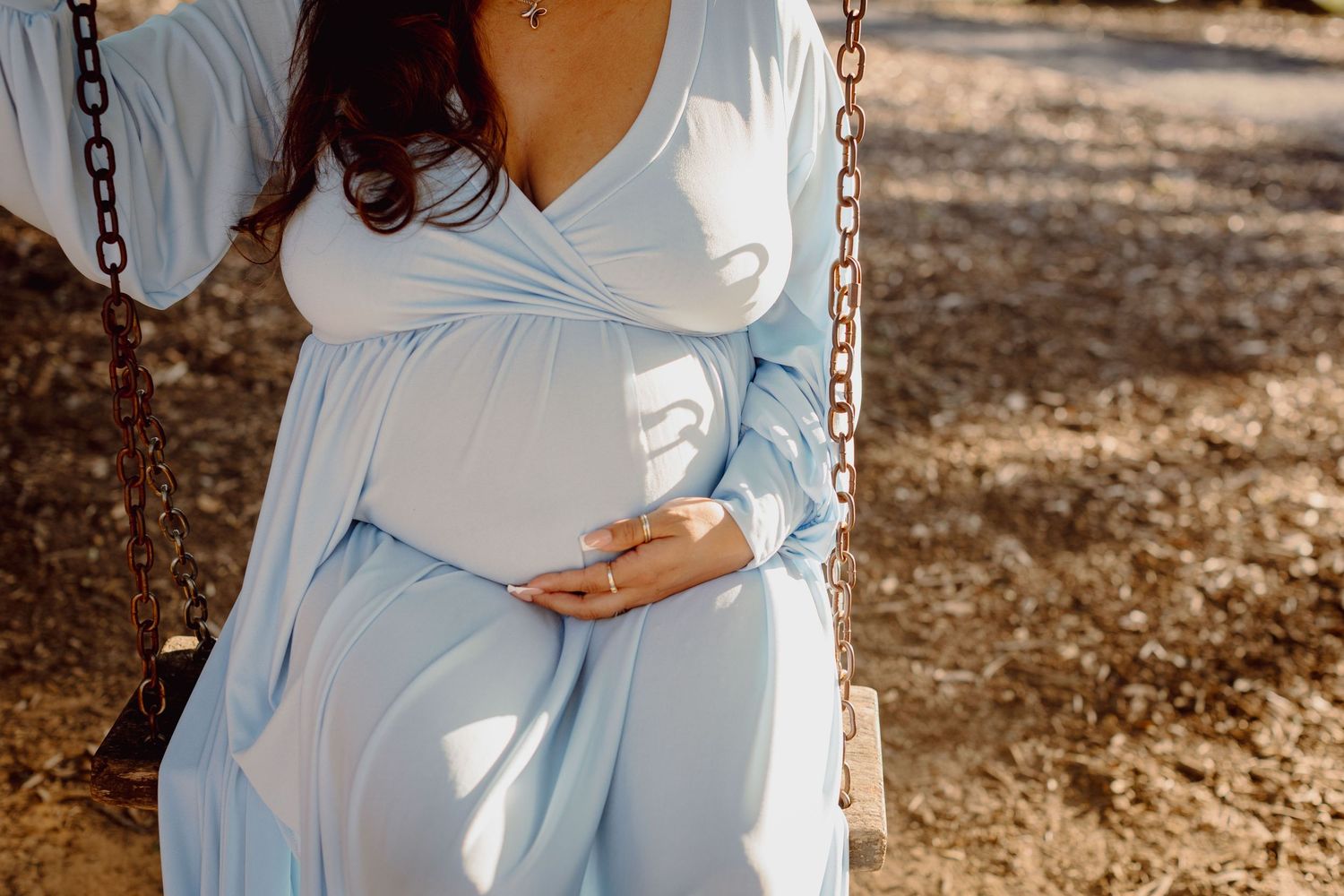 A maternity photoshoot in a white flowy dress shows someone cradling their baby bump in a sunlit outdoor setting.