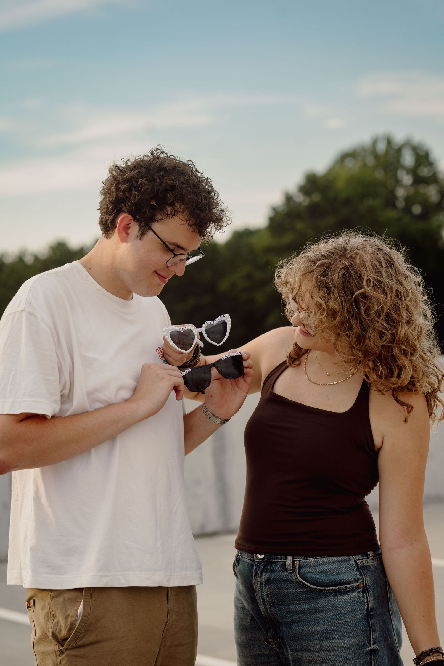 Young couple with curly hair examining heart-shaped sunglasses outdoors on a sunny day.