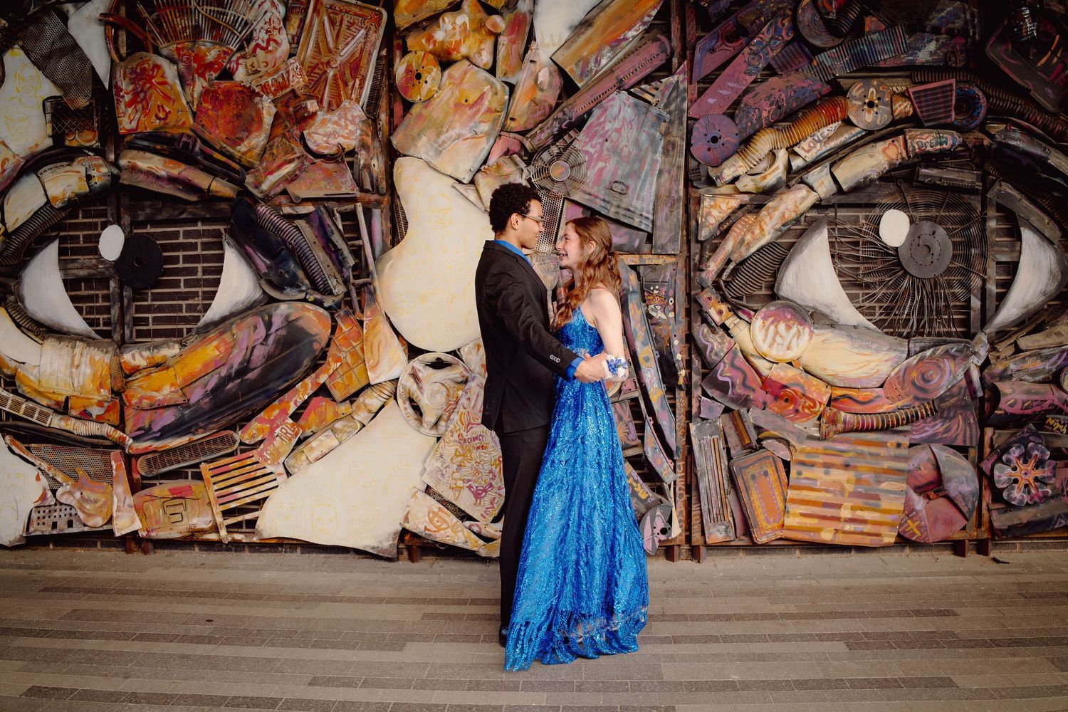 Couple in formal wear poses romantically before a large colorful street art mural featuring giant eyes.