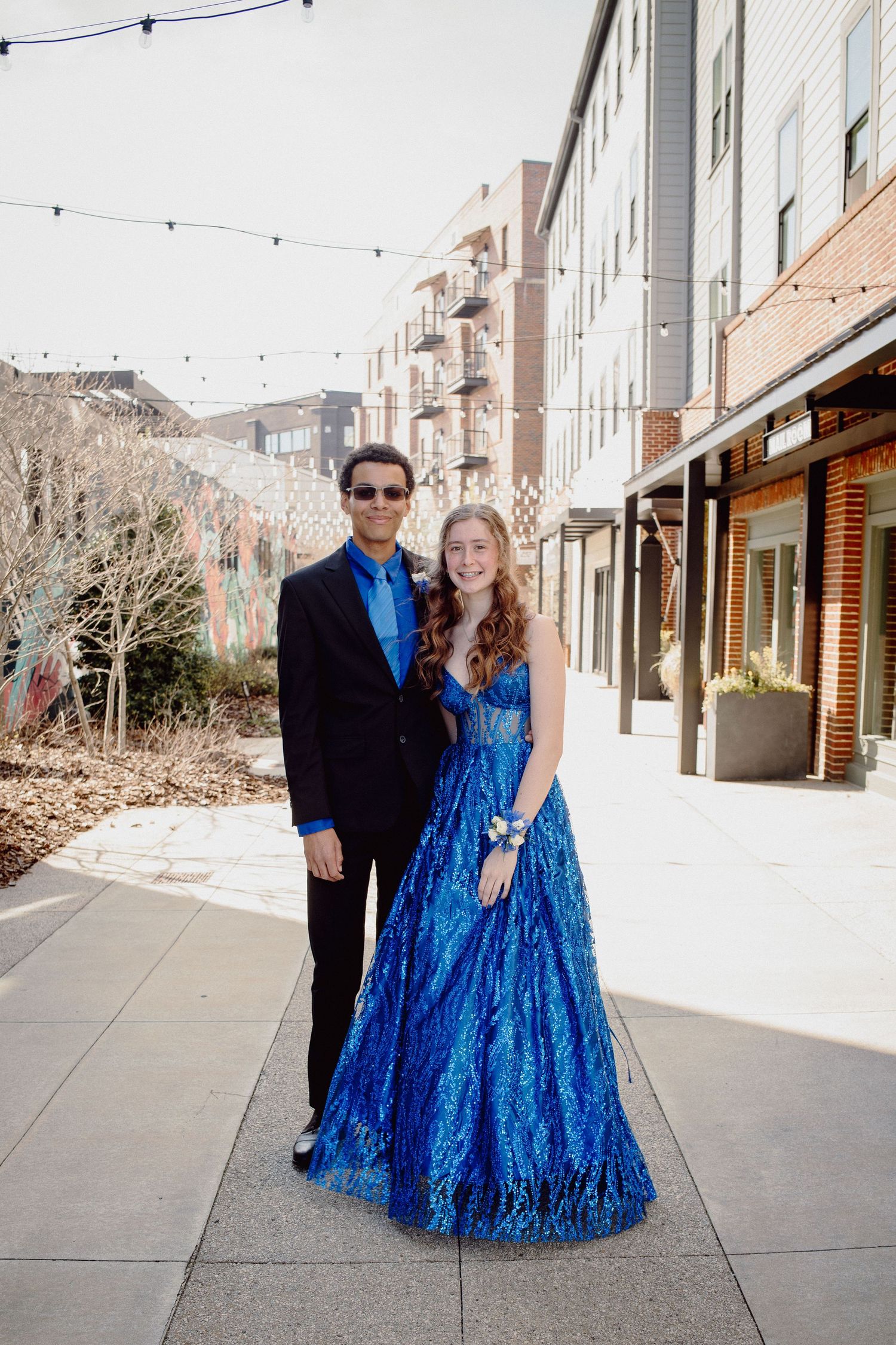 Couple in formal prom attire, girl in blue ball gown with corsage, boy in black suit, posing outdoors.
