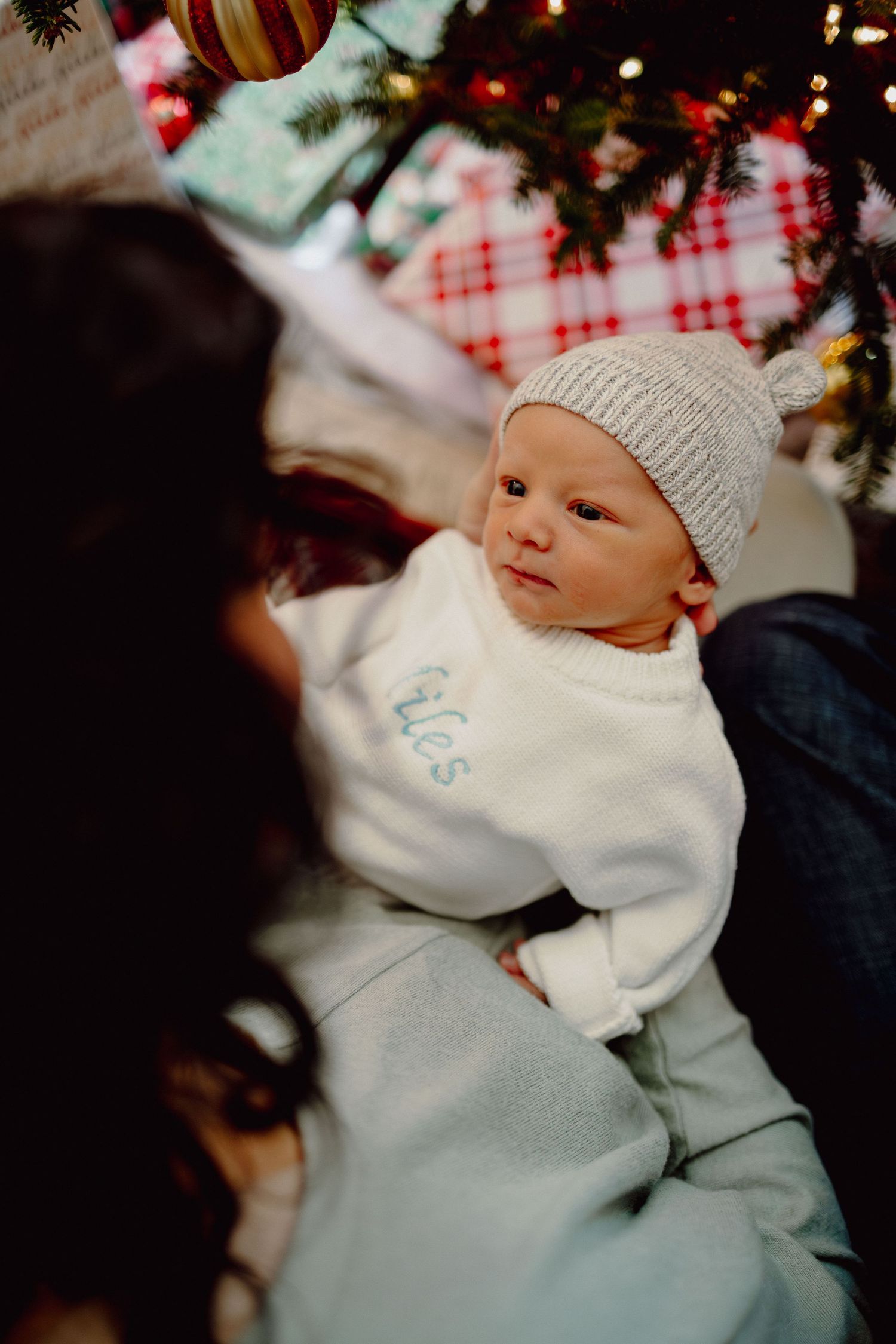 Baby wearing white knit hat and sweater lies under decorated Christmas tree with ornaments and lights.