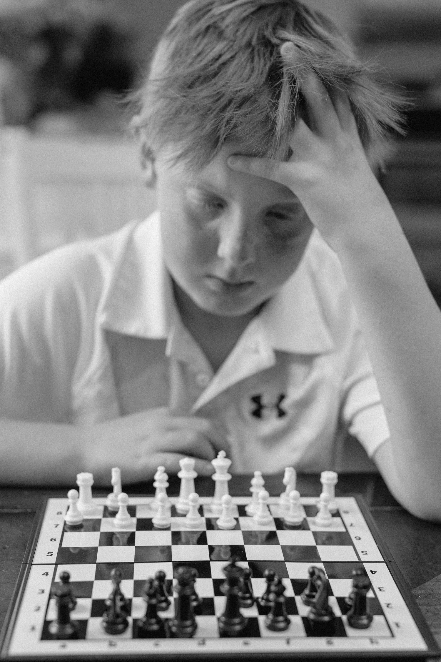 A young chess player in an Under Armour shirt contemplates their next move during a game in black and white photography.