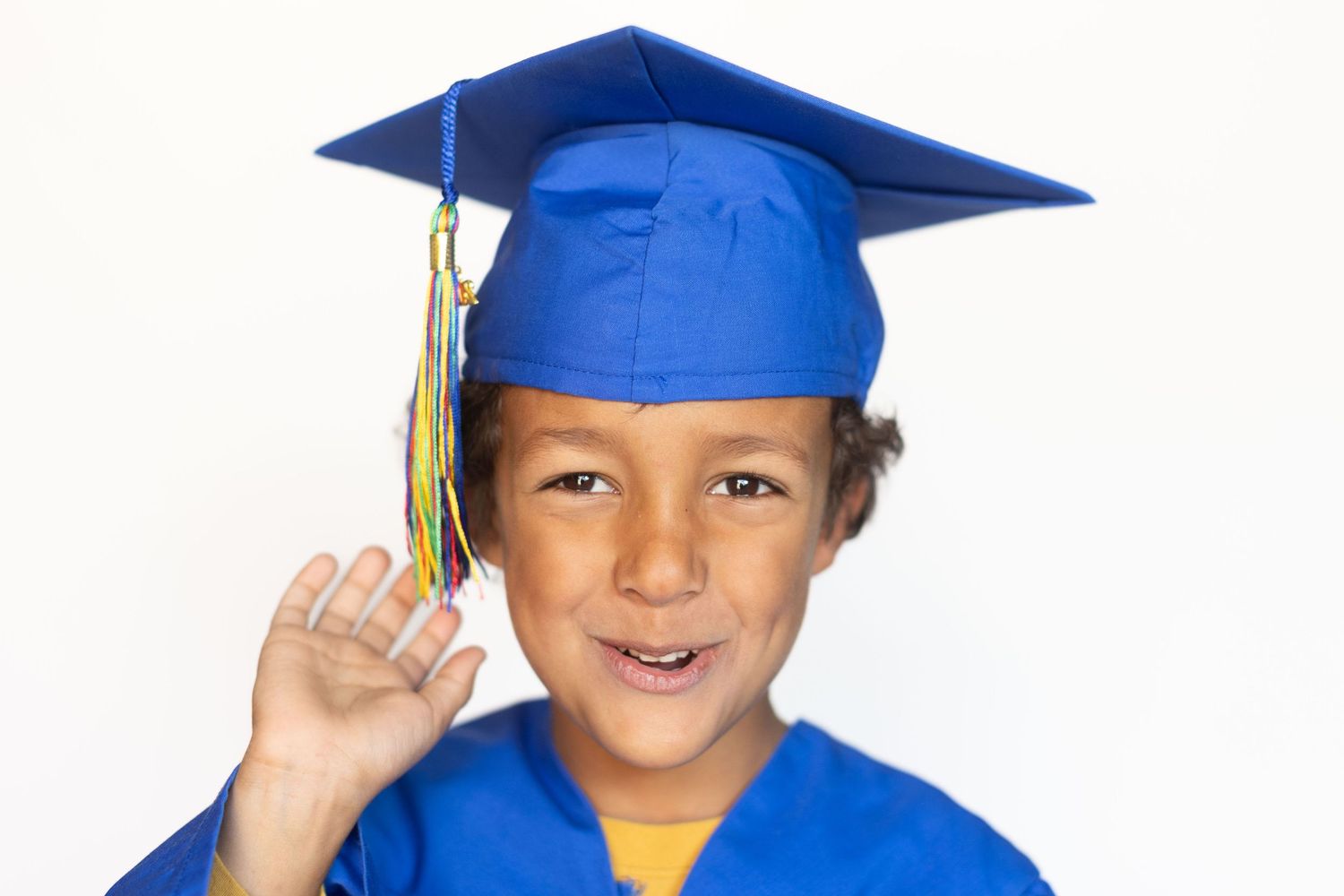 Young boy in blue graduation cap and gown smiling proudly, holding tassel on white background.