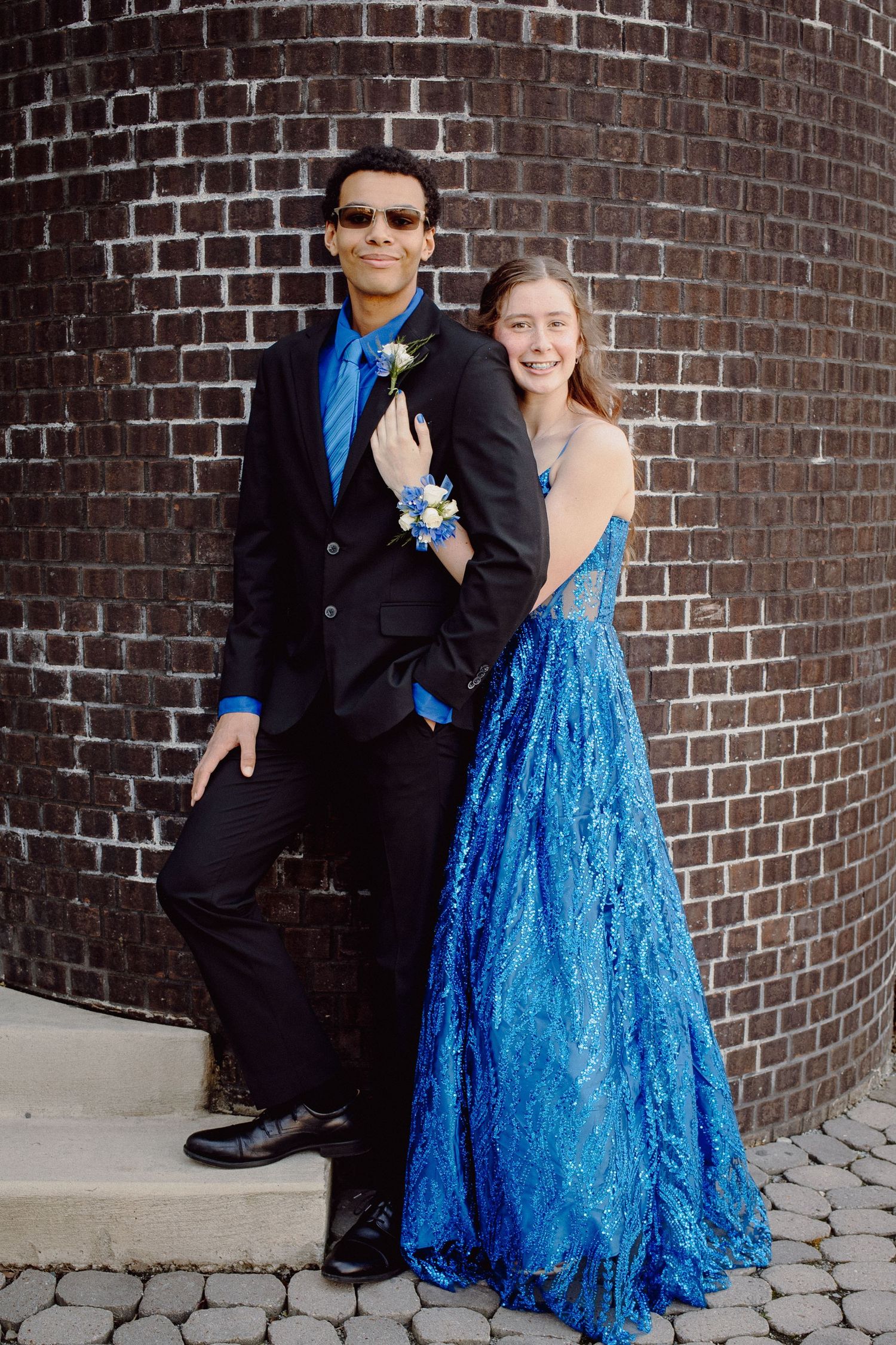 A young couple dressed in matching blue prom attire pose together in front of a brick wall.