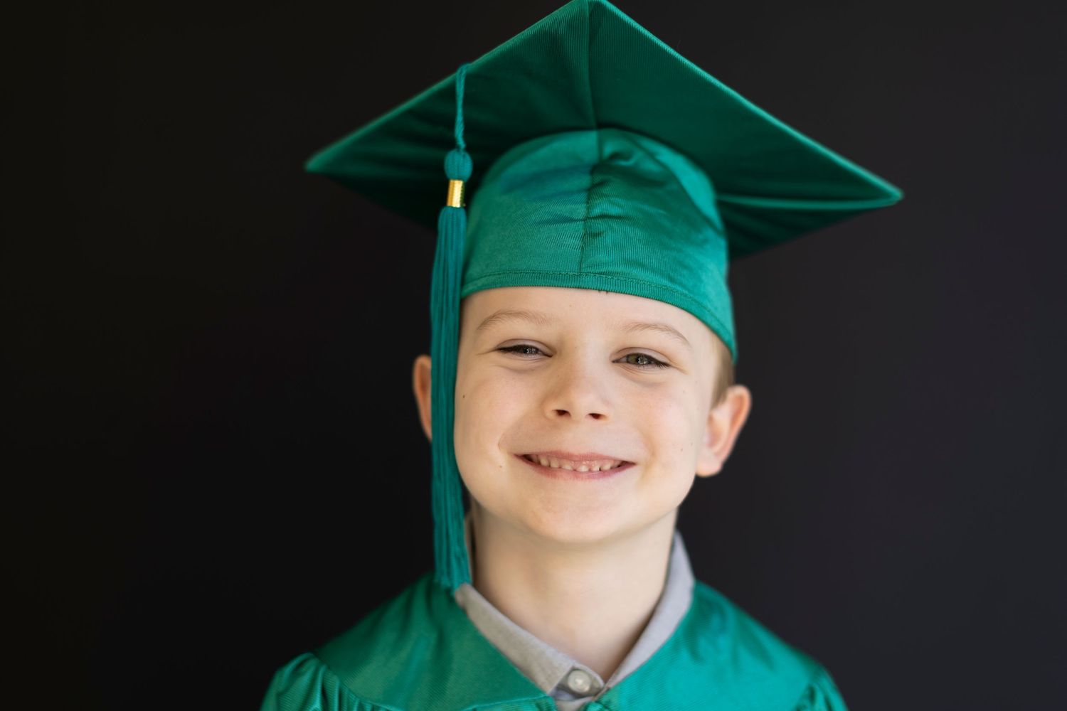 Smiling young boy wearing a green graduation cap and gown against a dark background.