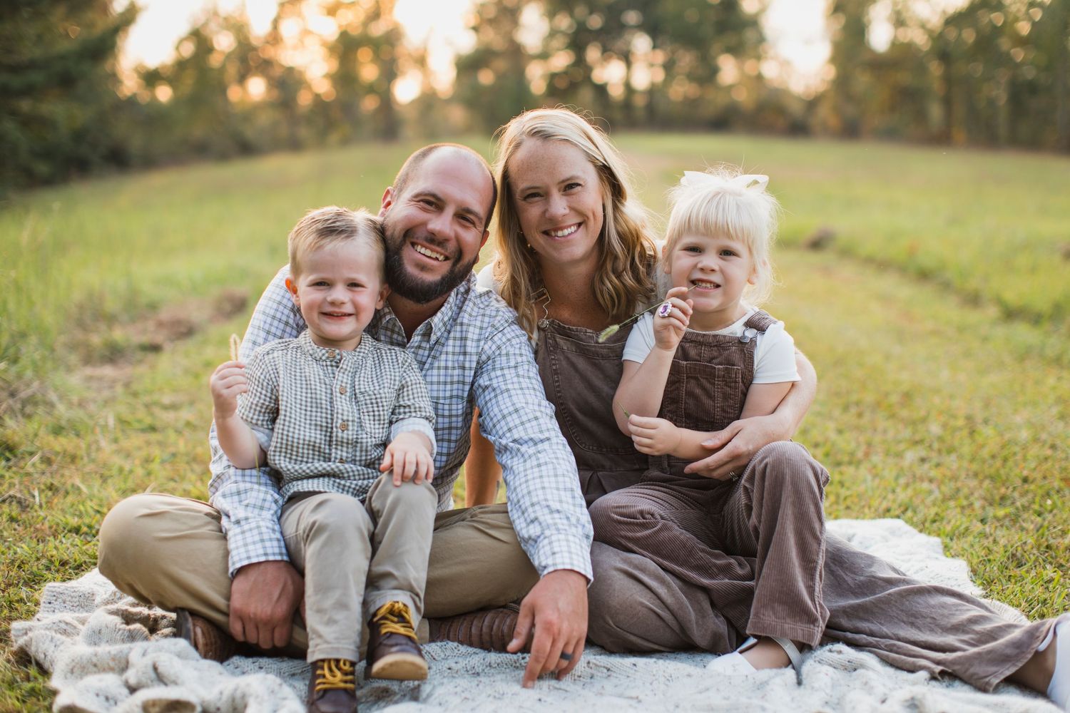 Happy family of four sitting on a blanket in a sunny green field, smiling together outdoors.