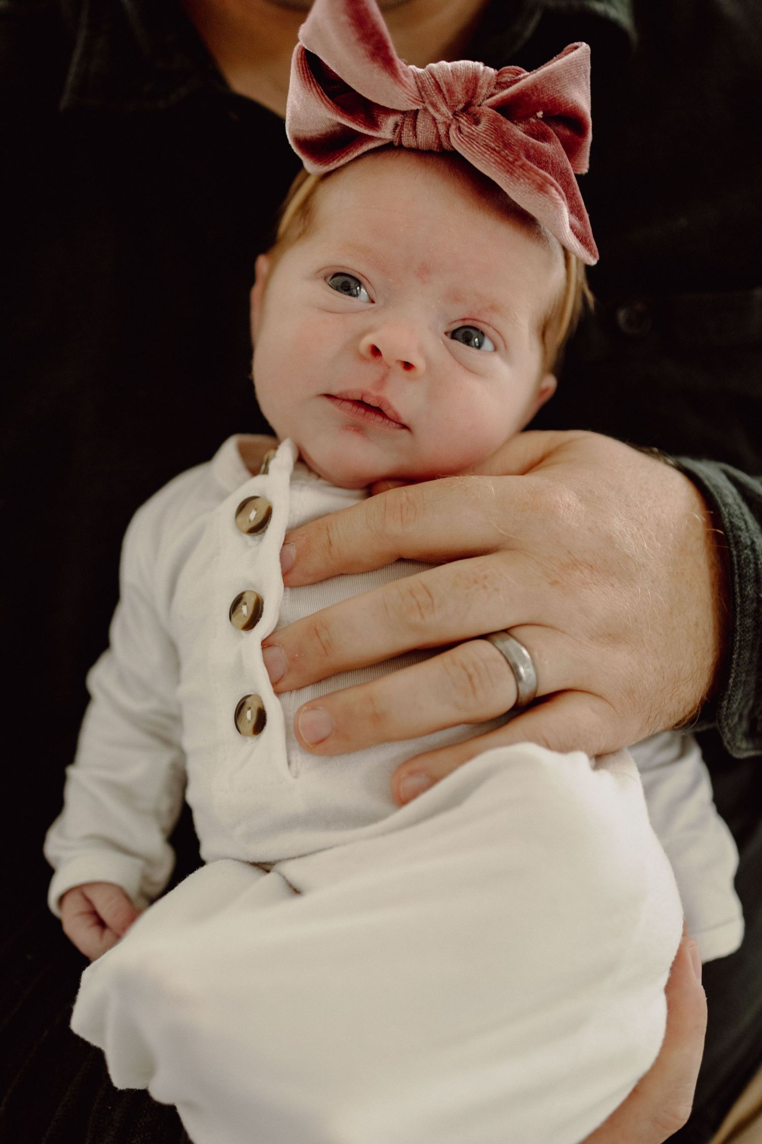 A baby wearing a white outfit and pink bow headband lies against a dark background.