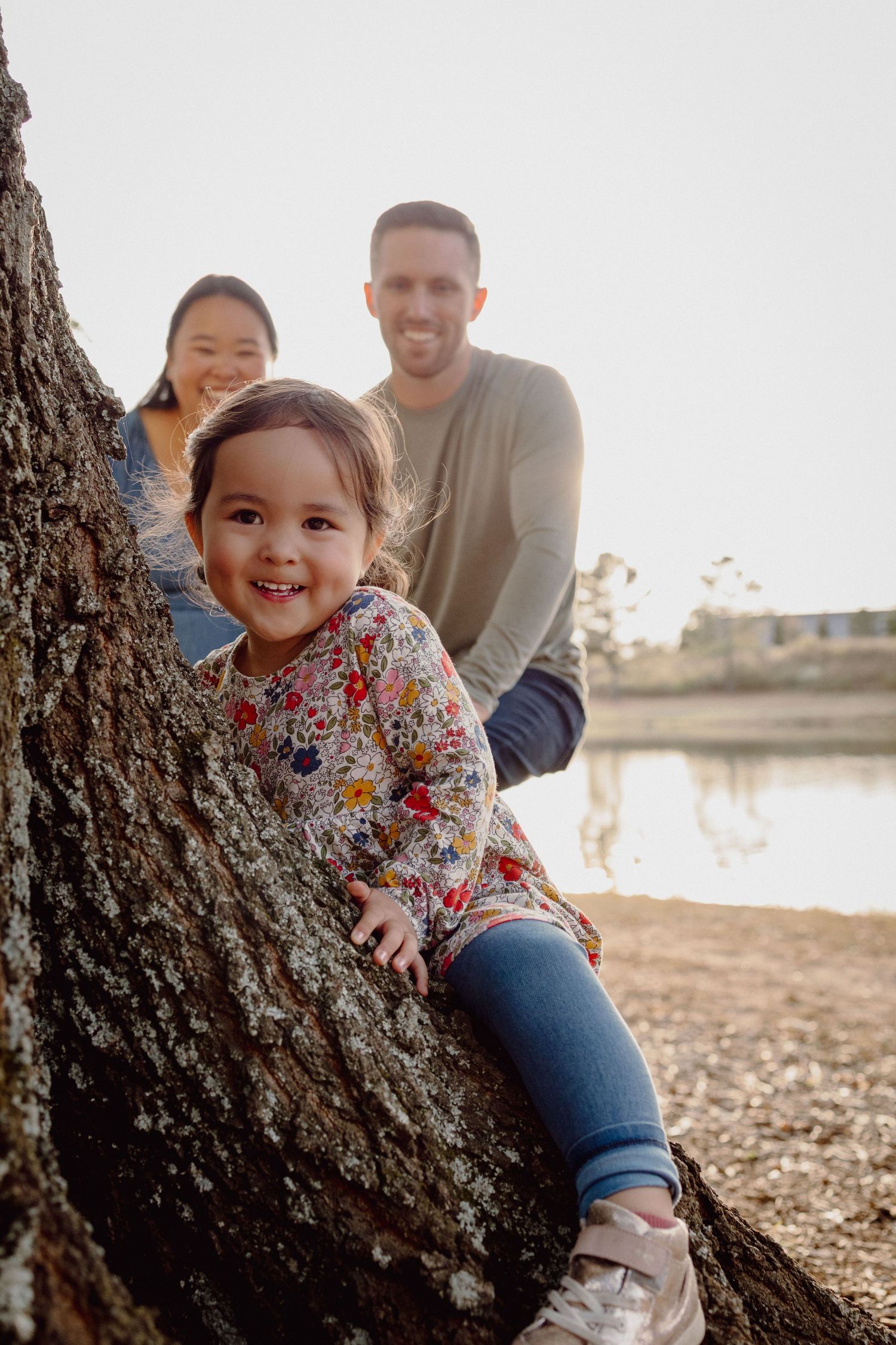 A family enjoys a sunny afternoon together by a tree near a lake, sharing smiles and laughter in nature.