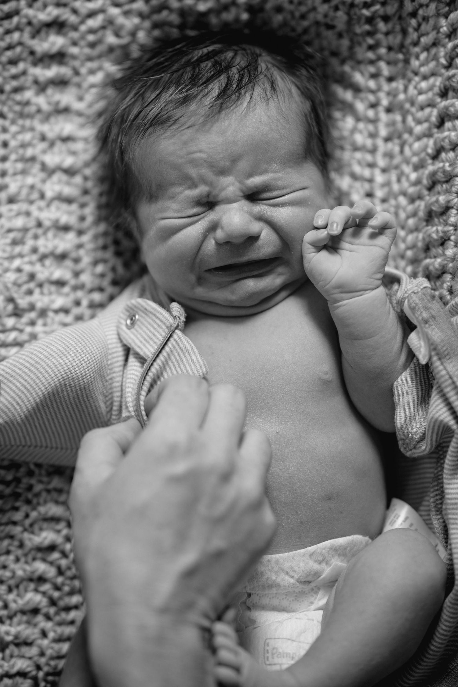 Black and white photo of a crying newborn baby in a diaper, lying on a knit blanket with an adult hand nearby.