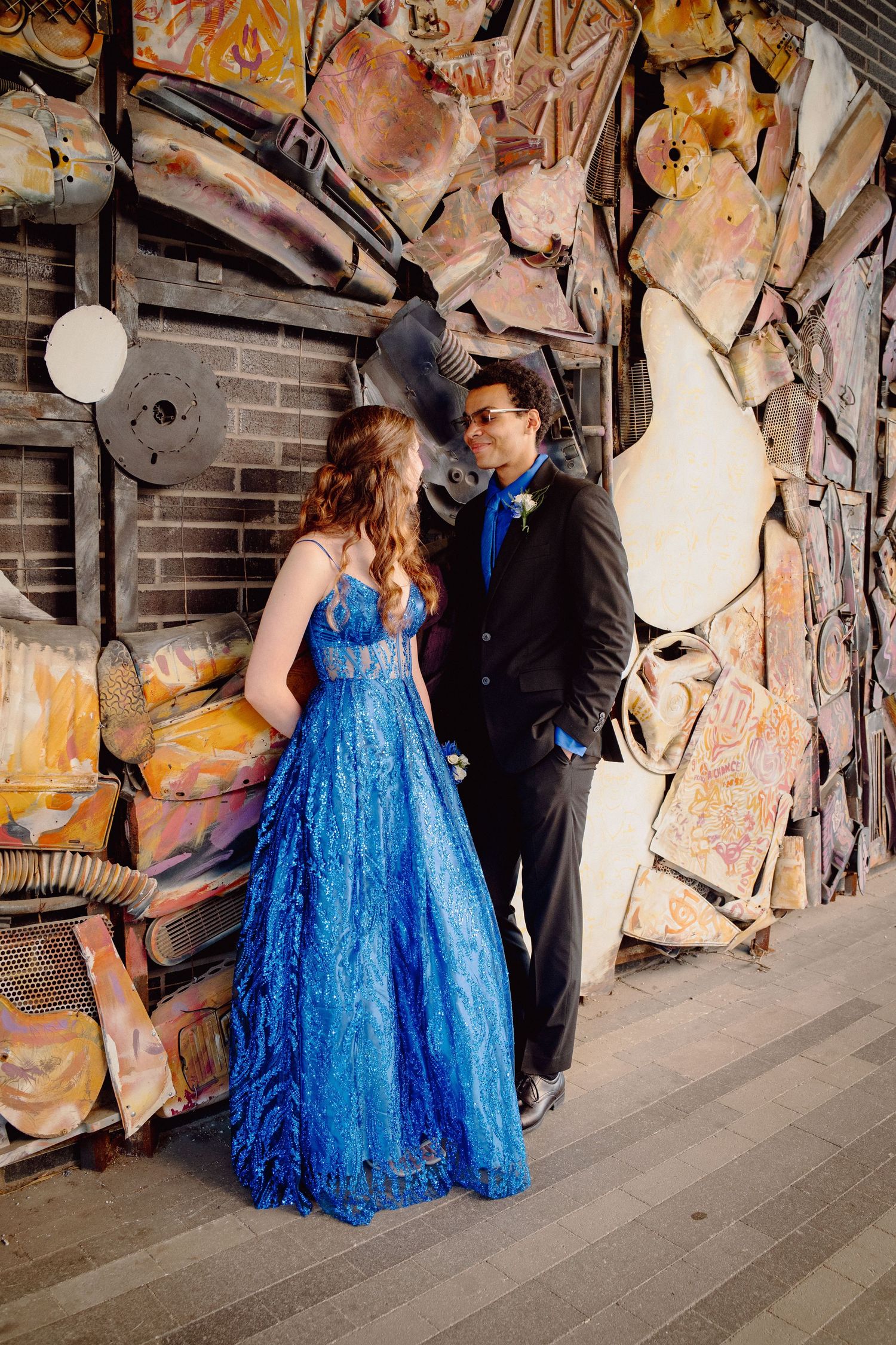 A couple in prom attire poses outdoors, the girl in a blue gown and the boy in a black suit with blue tie.