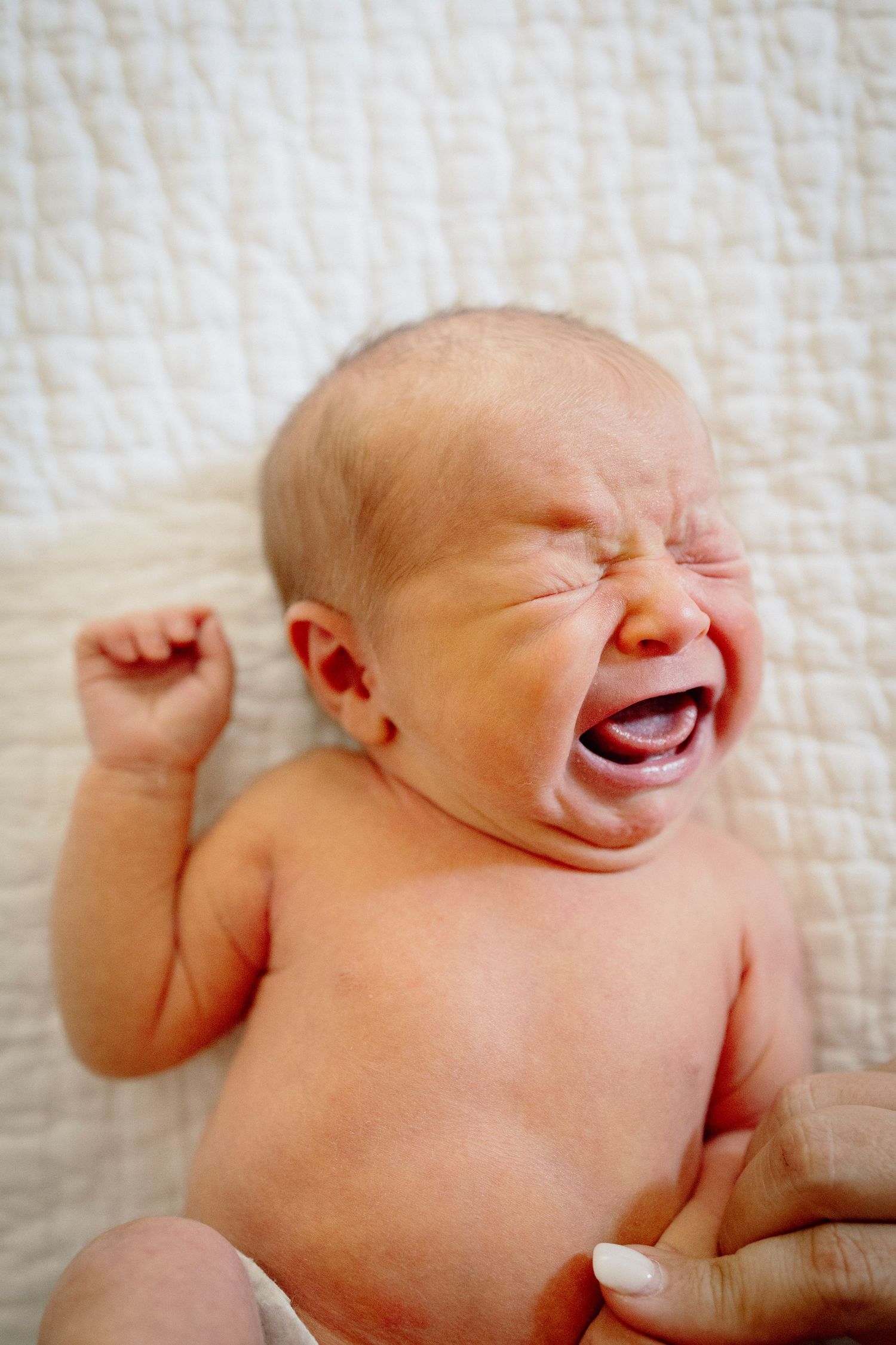 A happy baby lying on a white blanket laughs joyfully while stretching their arms.