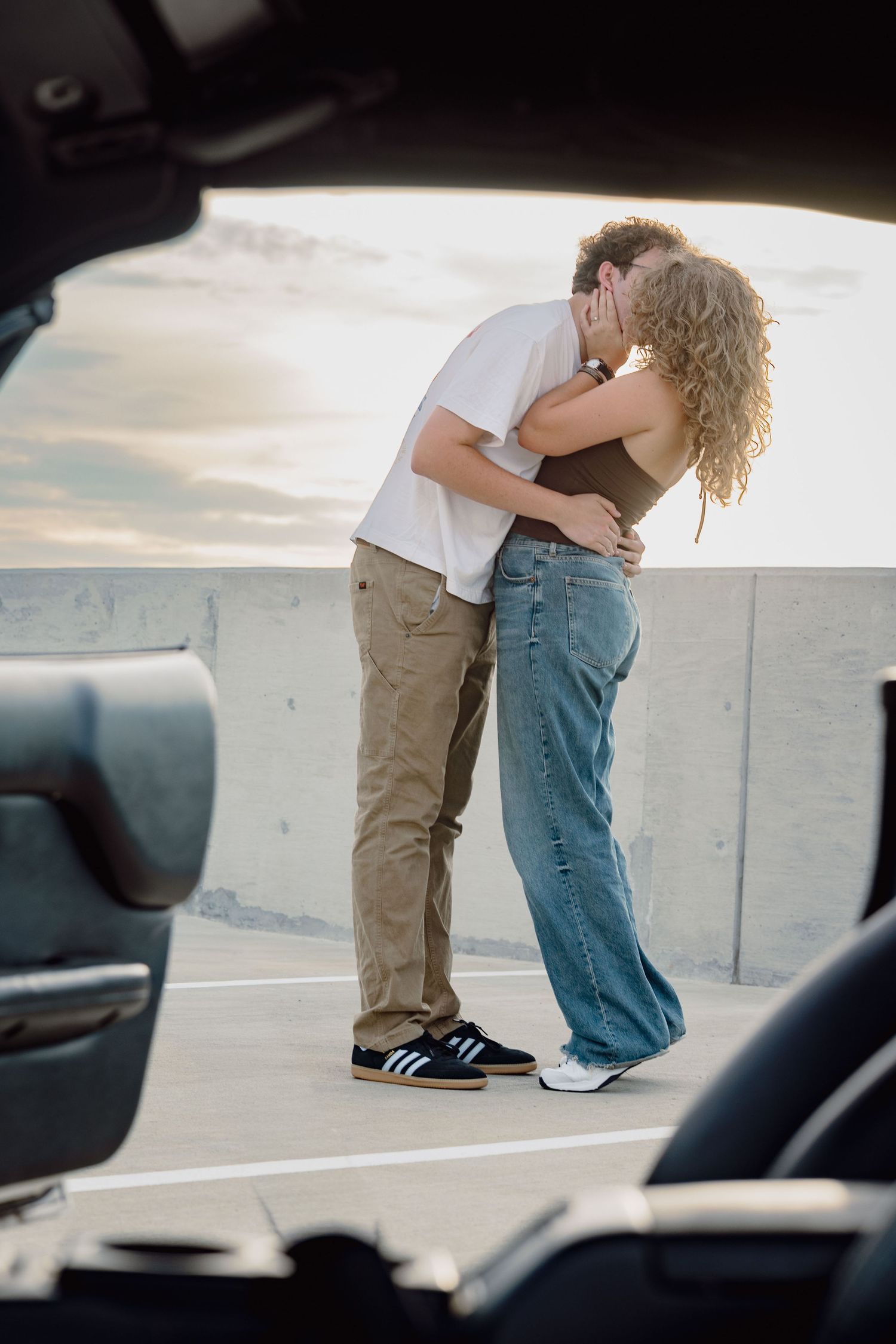 Couple sharing a romantic kiss on a rooftop parking lot at sunset, dressed in casual jeans and sneakers.