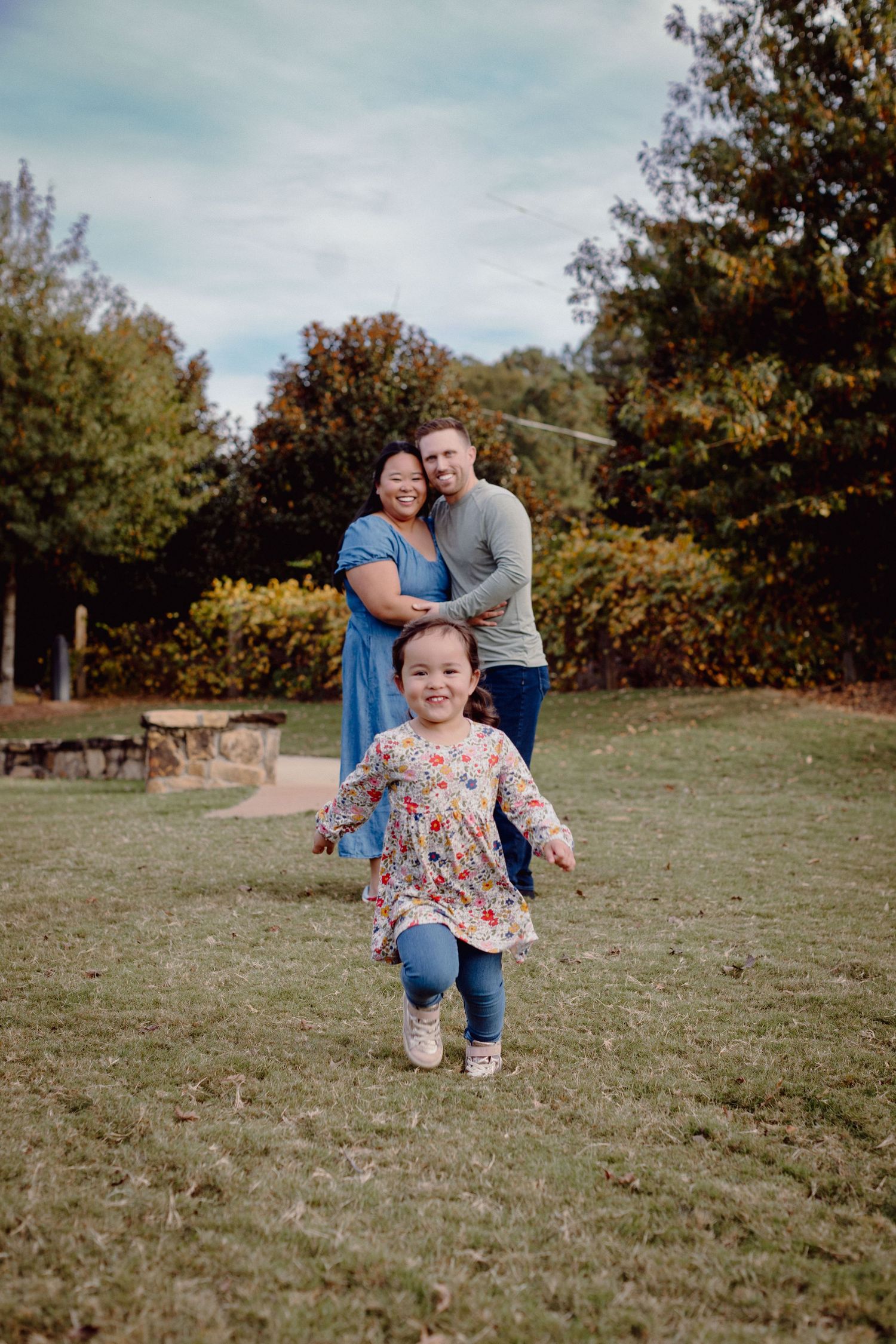 A happy family poses outdoors on green grass, with a young girl running forward and parents embracing behind her.