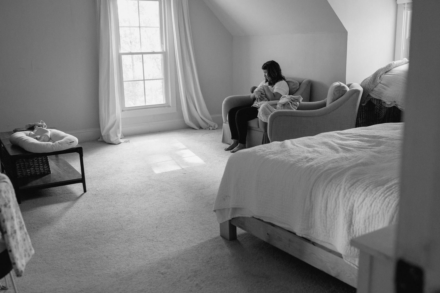 Black and white interior photograph showing a bright sunlit bedroom with minimalist decor and white bedding.