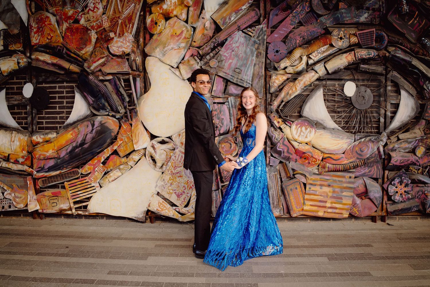 Couple in formal attire posing in front of a large colorful mural with eyes at a prom or gala event.