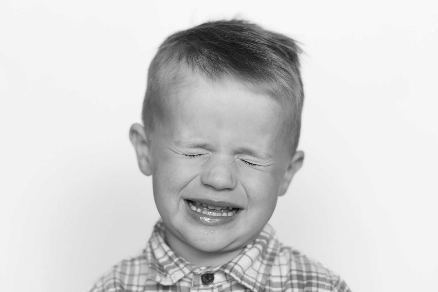Black and white portrait of a young toddler boy laughing with eyes closed, wearing a plaid shirt.