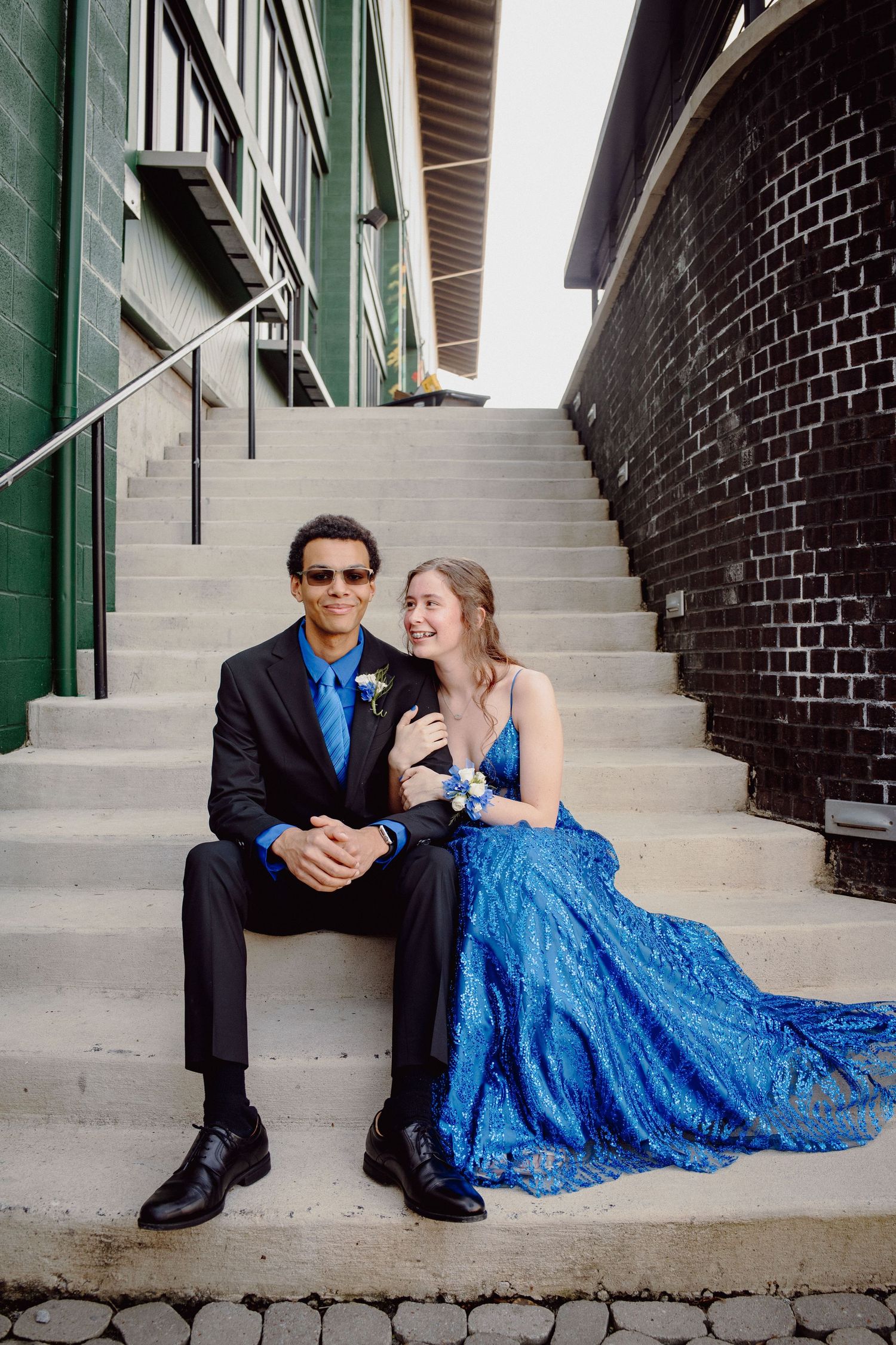 A couple in prom attire, man in black suit and woman in blue gown, sitting together on outdoor steps.